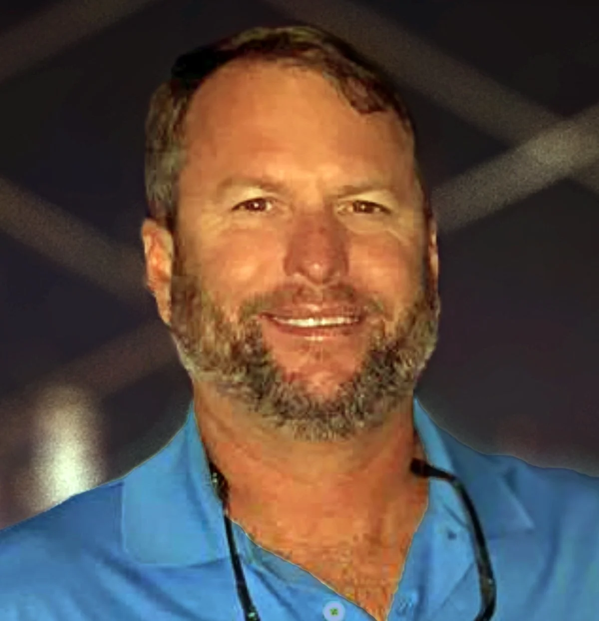 Portrait of a man with short hair and a beard, wearing a blue collared shirt, smiling at the camera.