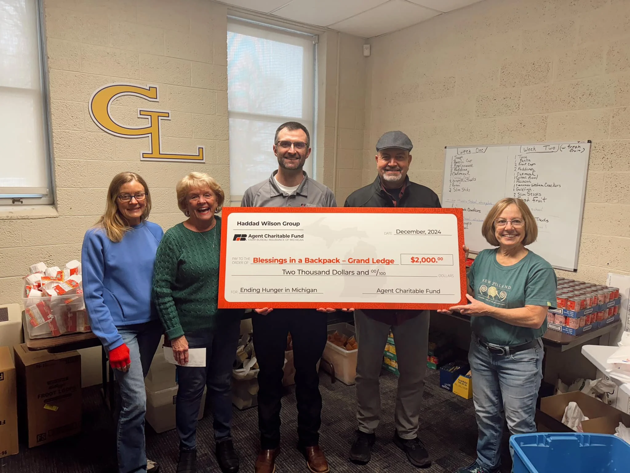 A group of five people standing indoors, holding a large check for $2,000 made out to Ending Hunger in Michigan, in an event organized by Haddad Wilson Group. Behind them are shelves with food items and a whiteboard with handwritten notes.