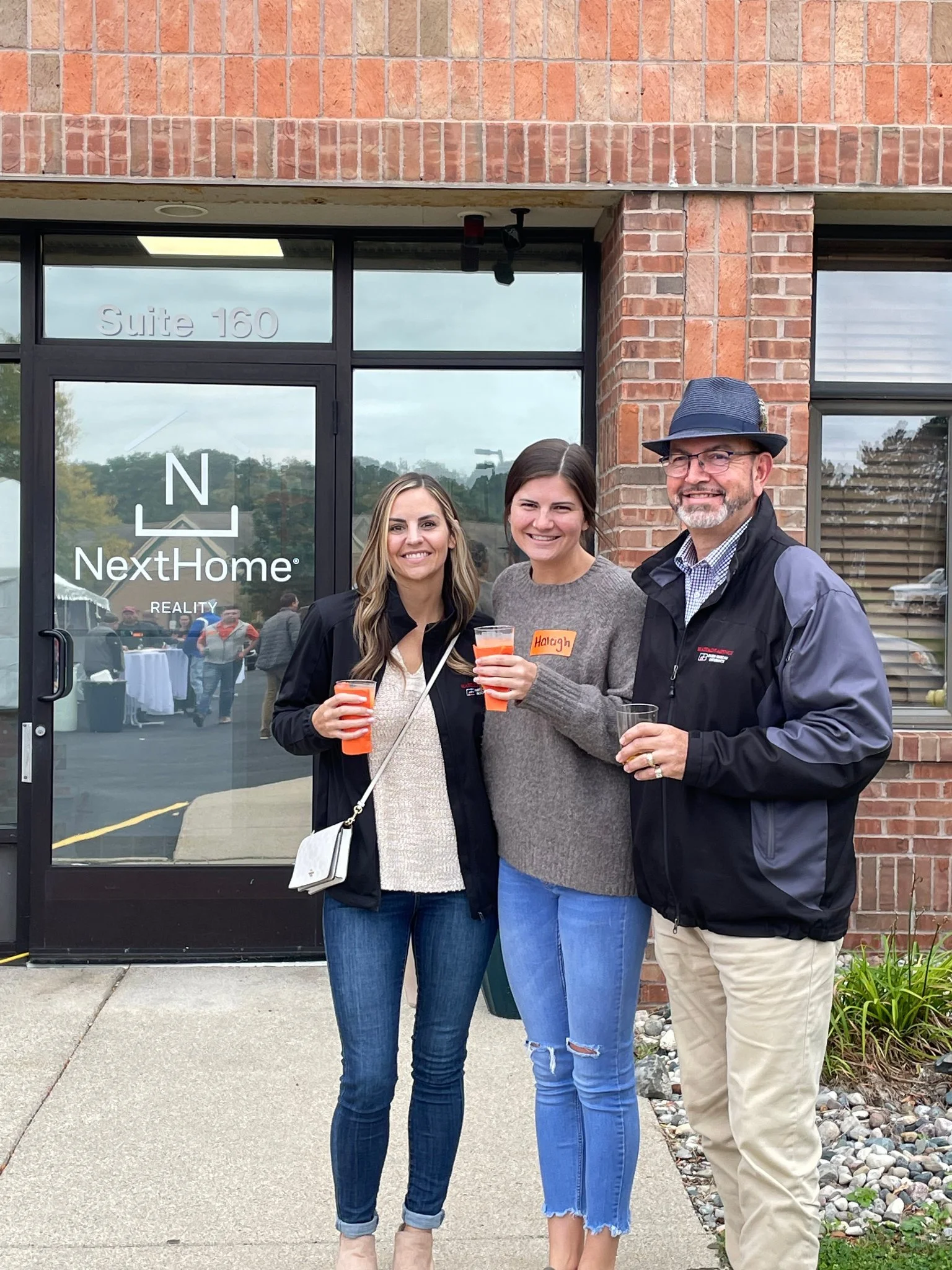Group of three people standing outside in front of a glass door with a logo for NextHome Real Estate. They are smiling and holding drinks. The woman on the left has long blonde hair, the woman in the middle has dark hair, and the man on the right wears a fedora hat and glasses.
