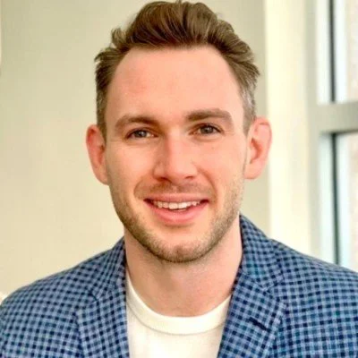 A smiling man with light skin, brown hair, and a beard, wearing a blue checkered blazer and white shirt, standing indoors near a window.