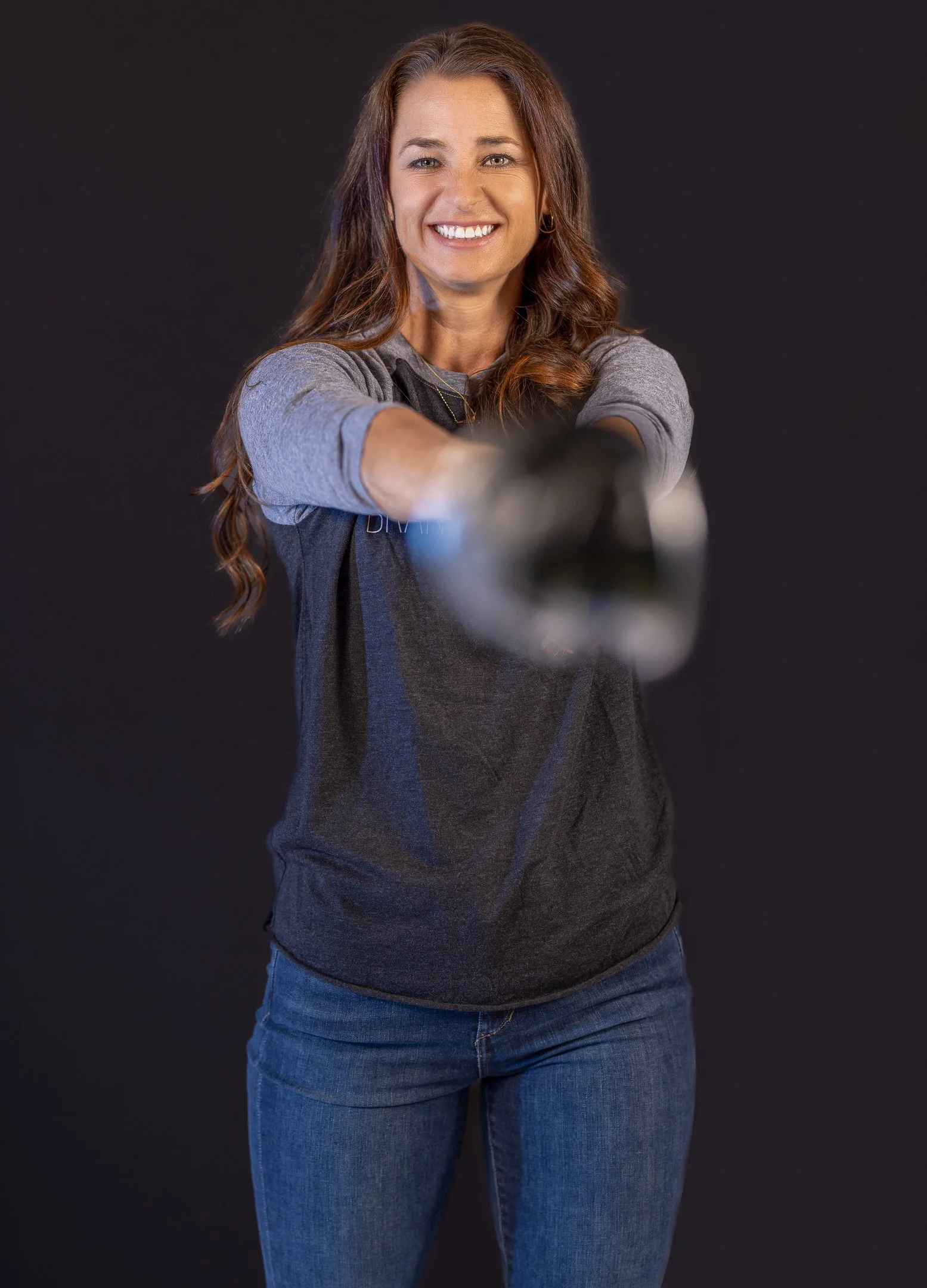 Woman with long brown hair smiling, extending her arm and pointing a finger, against a solid black background.