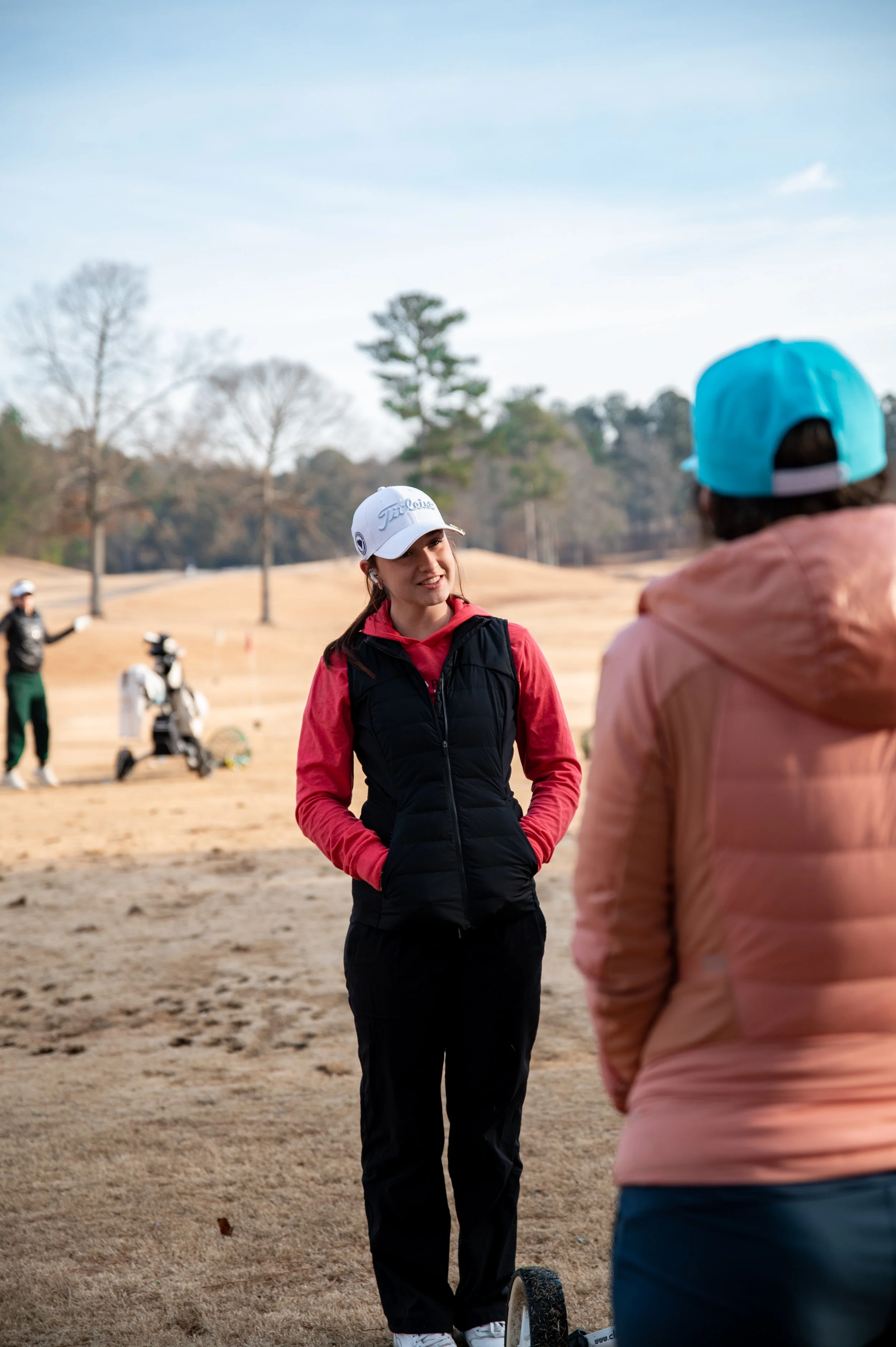 Two women talking on a golf course, one wearing a white cap, red hoodie, and black vest, the other in a pink jacket and blue cap, with golf carts and trees in the background.