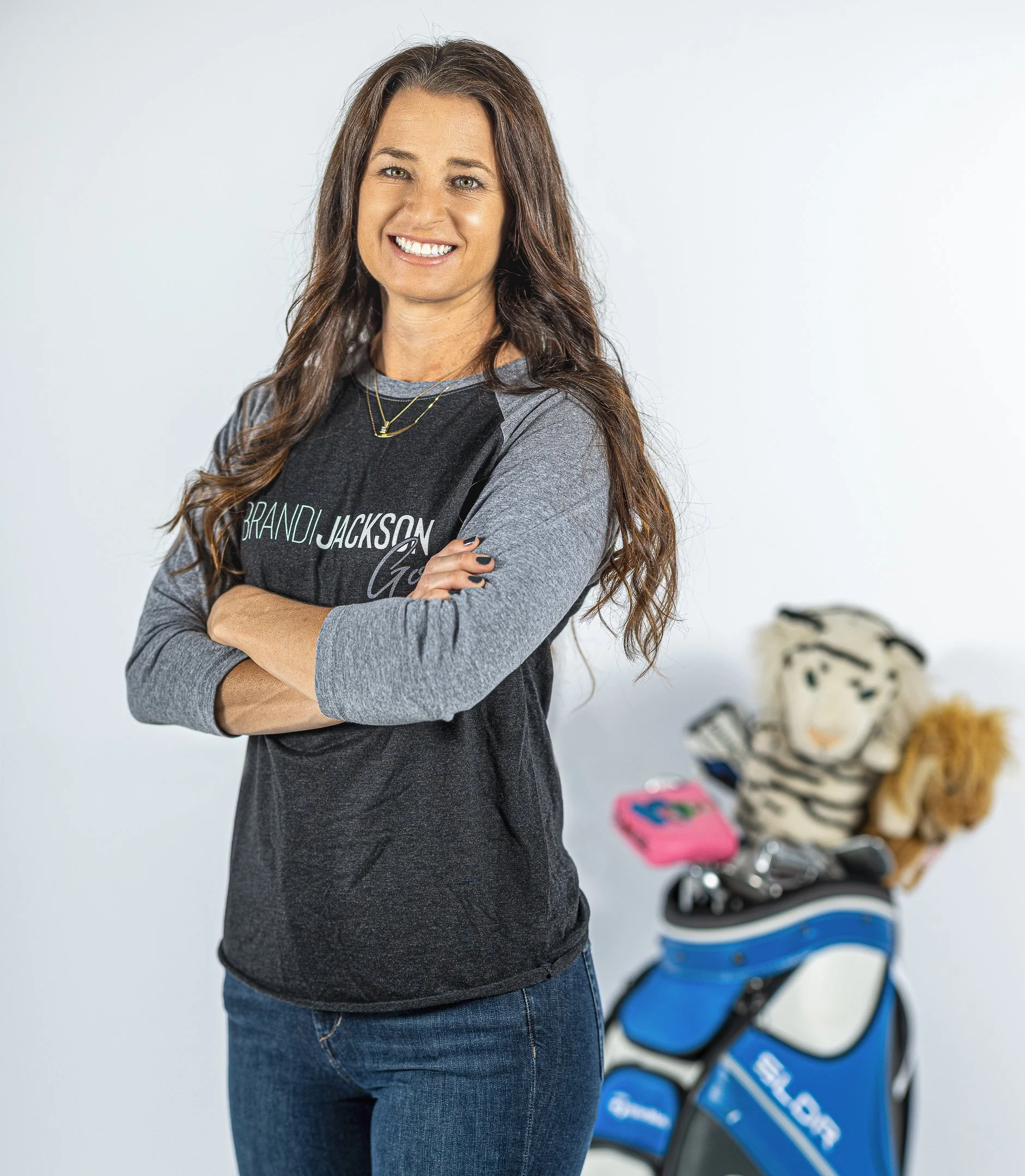 A woman with long brown hair smiling and crossing her arms, wearing a black and gray long-sleeve shirt, standing in front of a white background with a golf bag and stuffed tiger behind her.