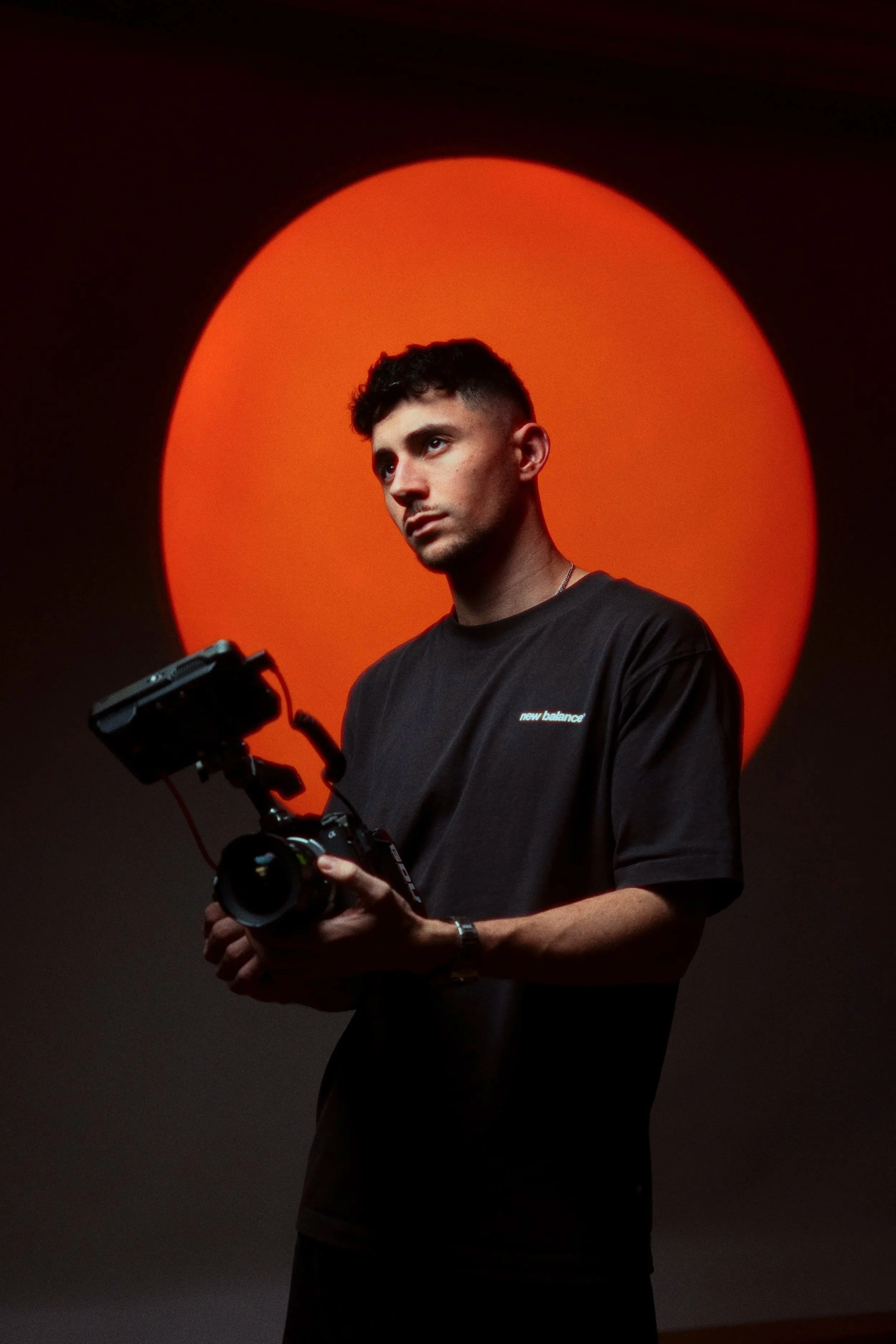 Junger Mann mit dunklem T-Shirt, das 'new balance' steht, hält eine Kamera in den Händen und schaut nach oben, vor einem großen roten Halbkreis.
