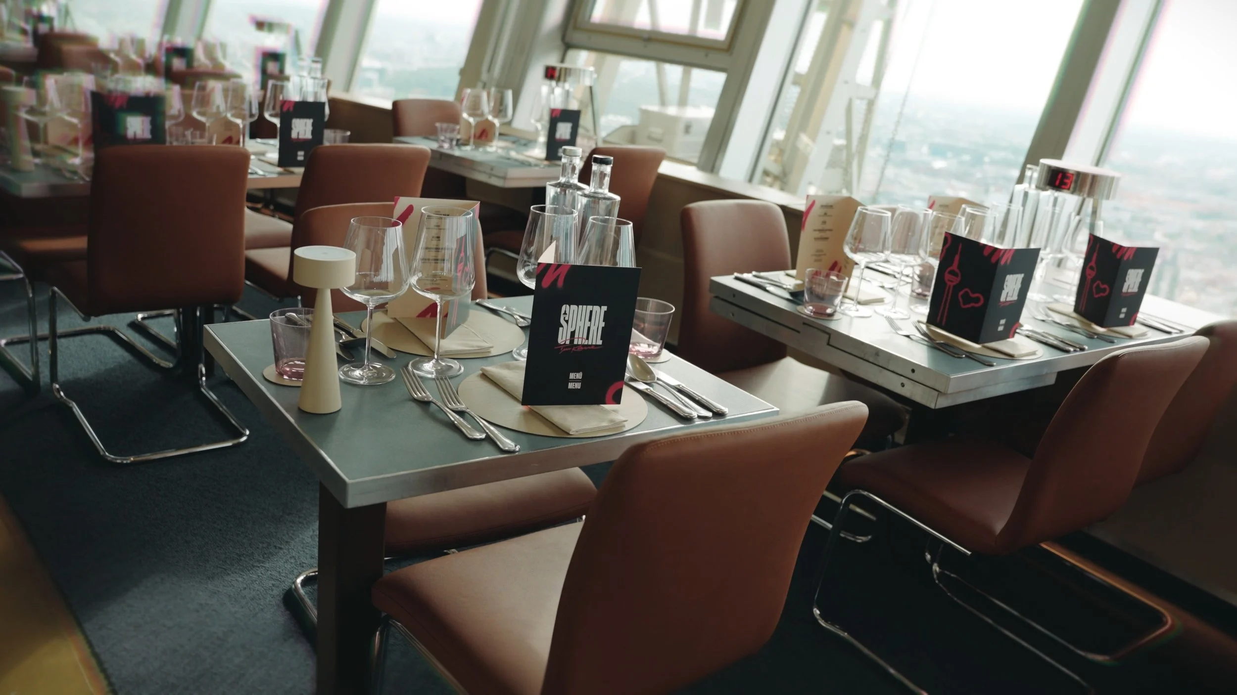 Ein stilvoll gedeckter Esstisch in einem modernen Hochhausrestaurant mit Blick auf die Stadt, mit Wein- und Wassergläsern, Besteck, Menükarten und einem kleinen Tischläufer.
