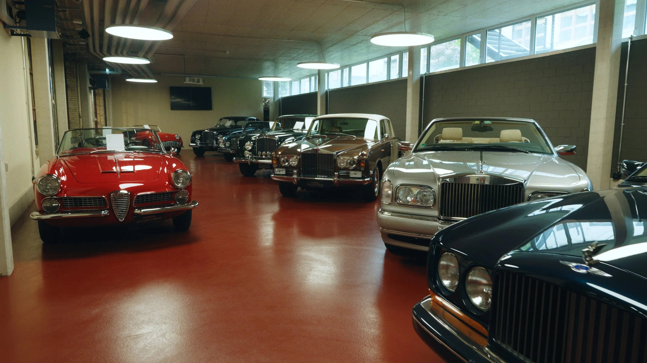 Sammlung von klassischen Autos in einem Raum mit glatten roten Boden und großen Fenstern an der Decke.