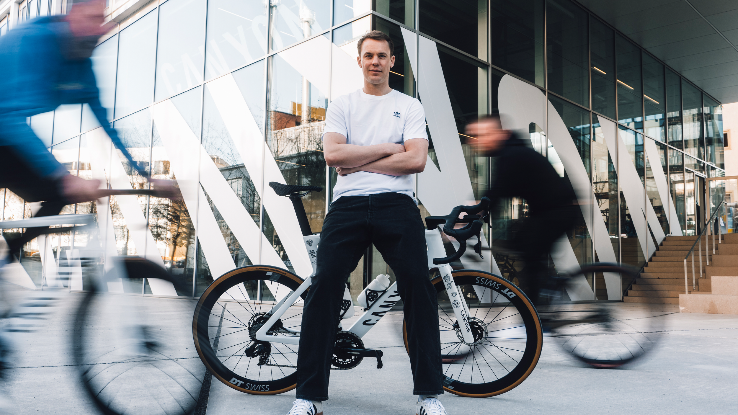 Junger Mann steht mit verschränkten Armen neben einem Fahrrad vor einem modernen Glasgebäude, während Radfahrer vorbeifahren.