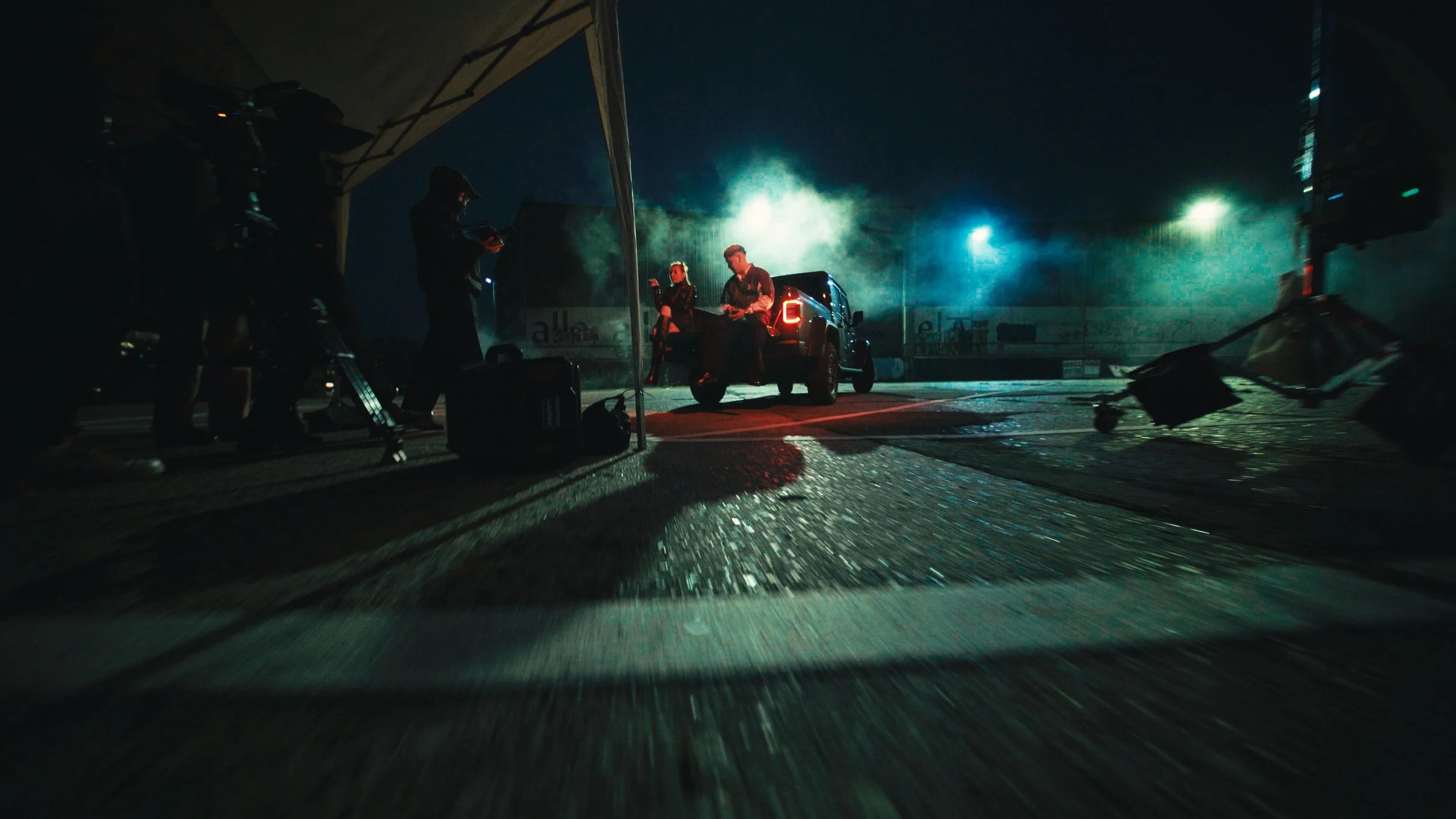 Dreharbeiten bei Nacht auf einer Rennstrecke, Kamera und Equipment sind sichtbar, zwei Personen sitzen im Heck eines schwarzen Autos, während andere im Schatten arbeiten.