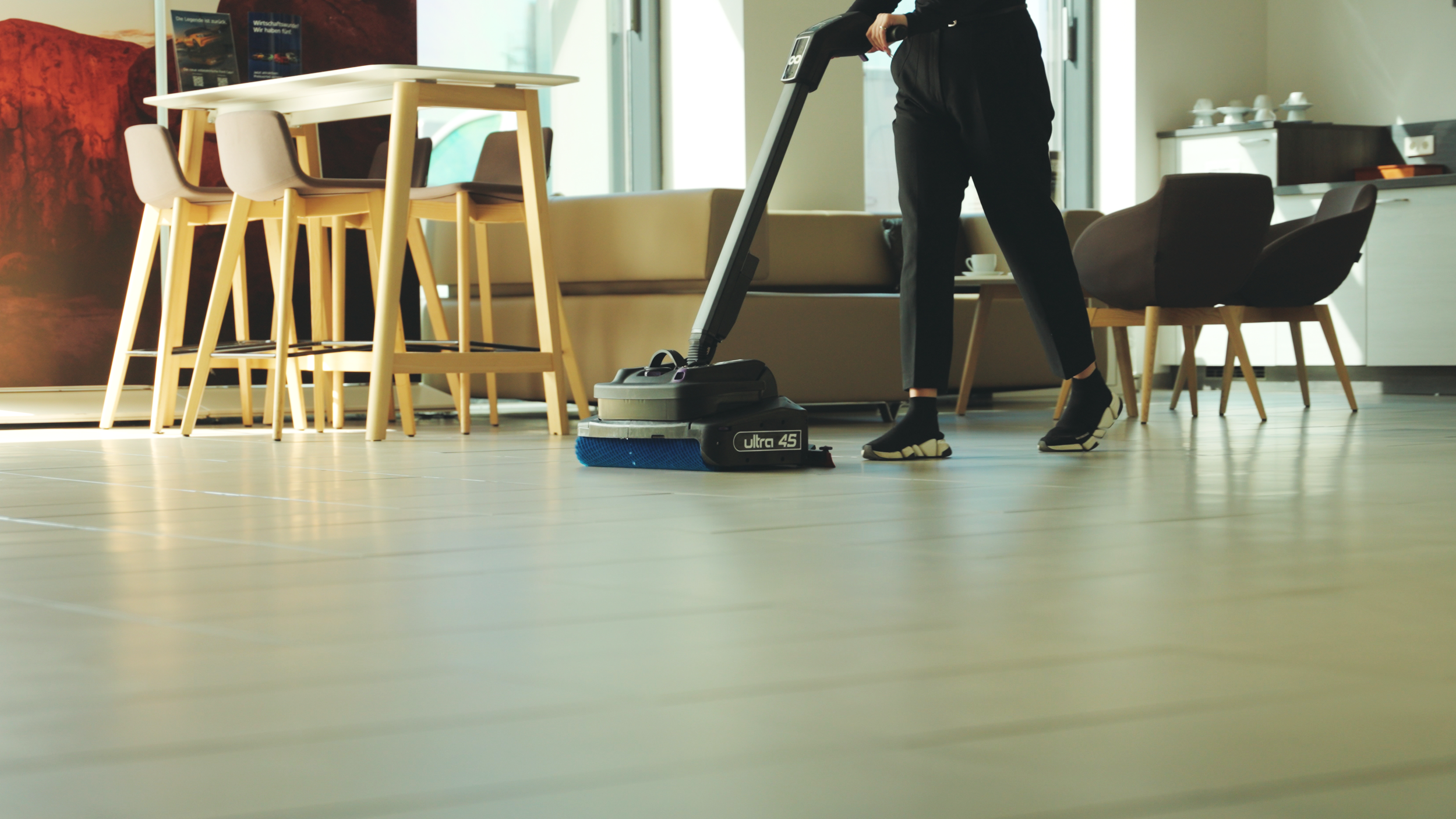 Person reinigt den Boden mit einem elektrischen Staubsauger in einem modernen Wohnzimmer mit Esstisch, Stühlen und Sitzgelegenheiten.