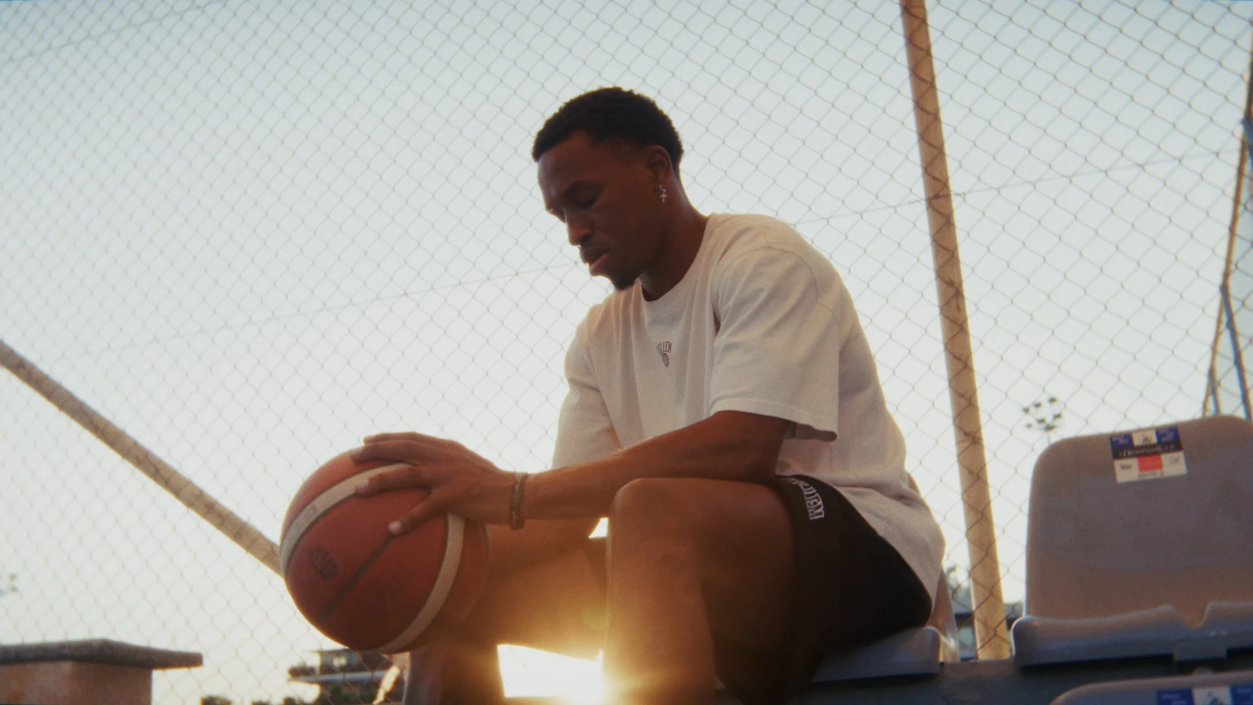 Junger Mann sitzt auf einer Bank, hält einen Basketball, trägt ein T-Shirt und Shorts, im Hintergrund ein Kletternetz und eine Tennisplatte, symbolisiert Freizeit im Freien.