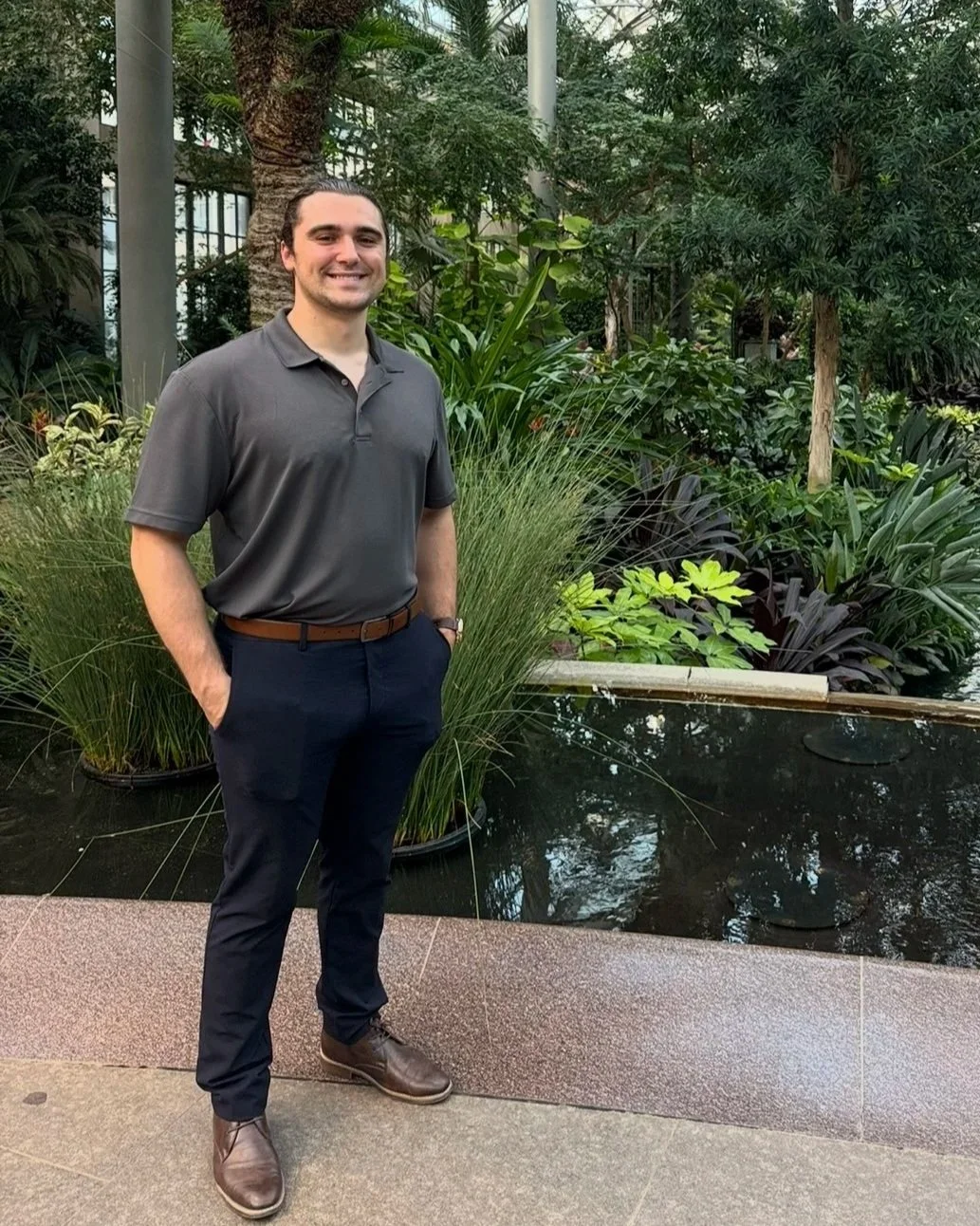 A man standing outdoors in a garden or atrium with lush green plants and trees in the background, wearing a gray polo shirt, dark pants, and brown shoes, smiling with his hands in his pockets.