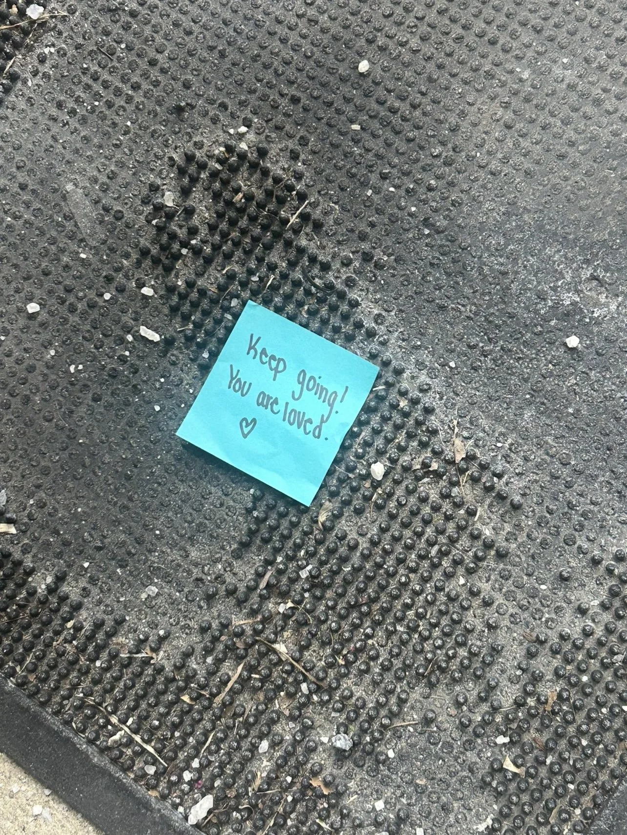 A blue sticky note with the message 'Keep going! You are loved.' and a small heart symbol, placed on a textured outdoor surface with dirt, small rocks, and debris around it.