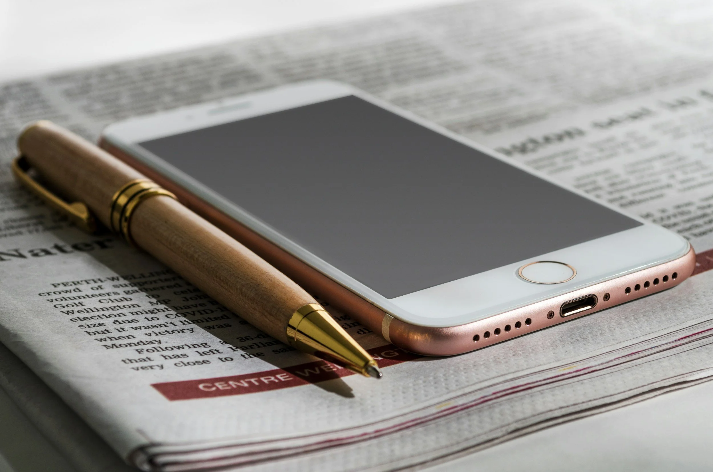Close-up of a smartphone with a rose gold border and white front, placed on top of a newspaper. A wooden pen with gold accents lies beside the phone on the newspaper.