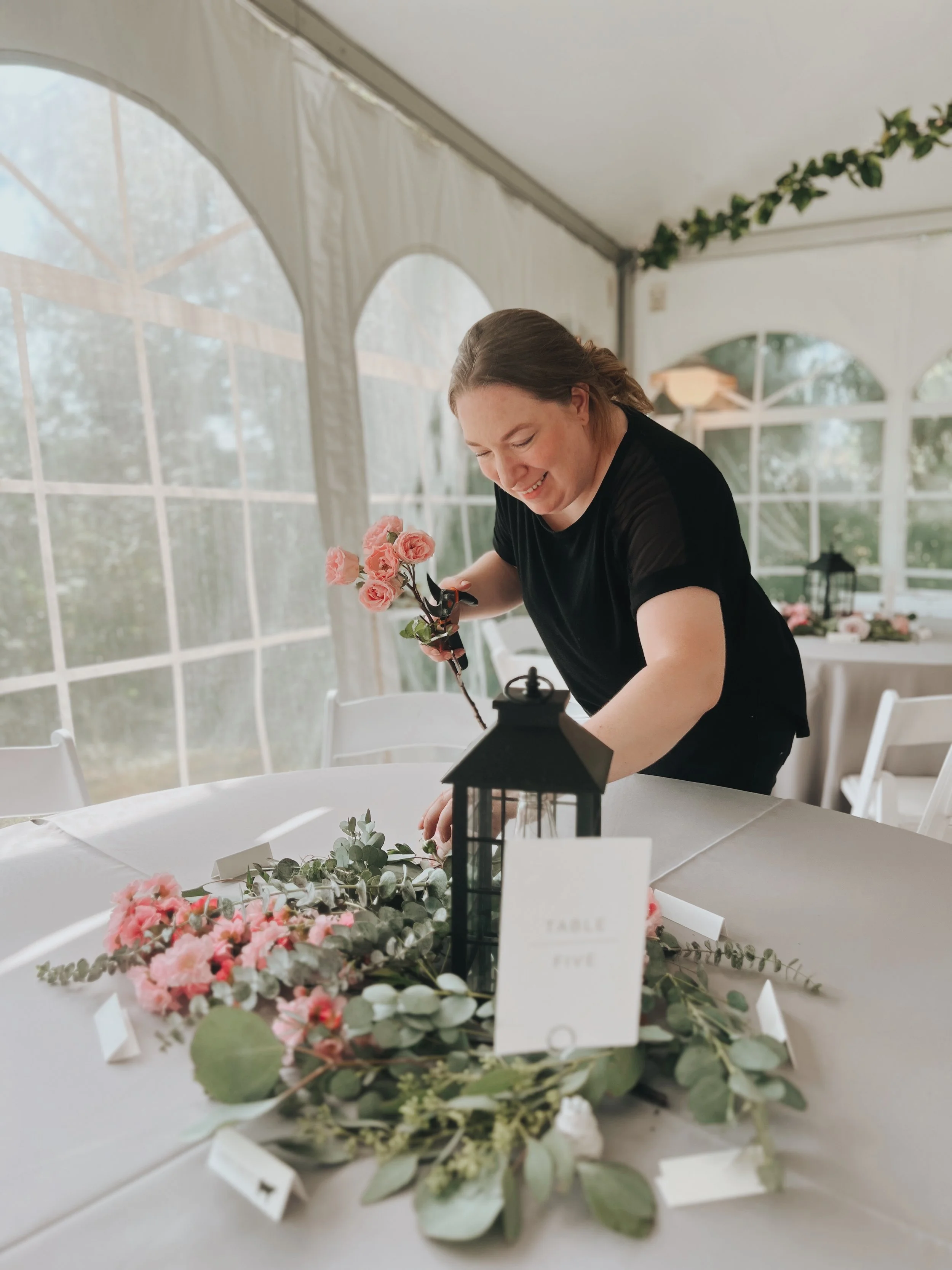 Woman decorating a wedding or event table with pink flowers and greenery inside a bright tent.