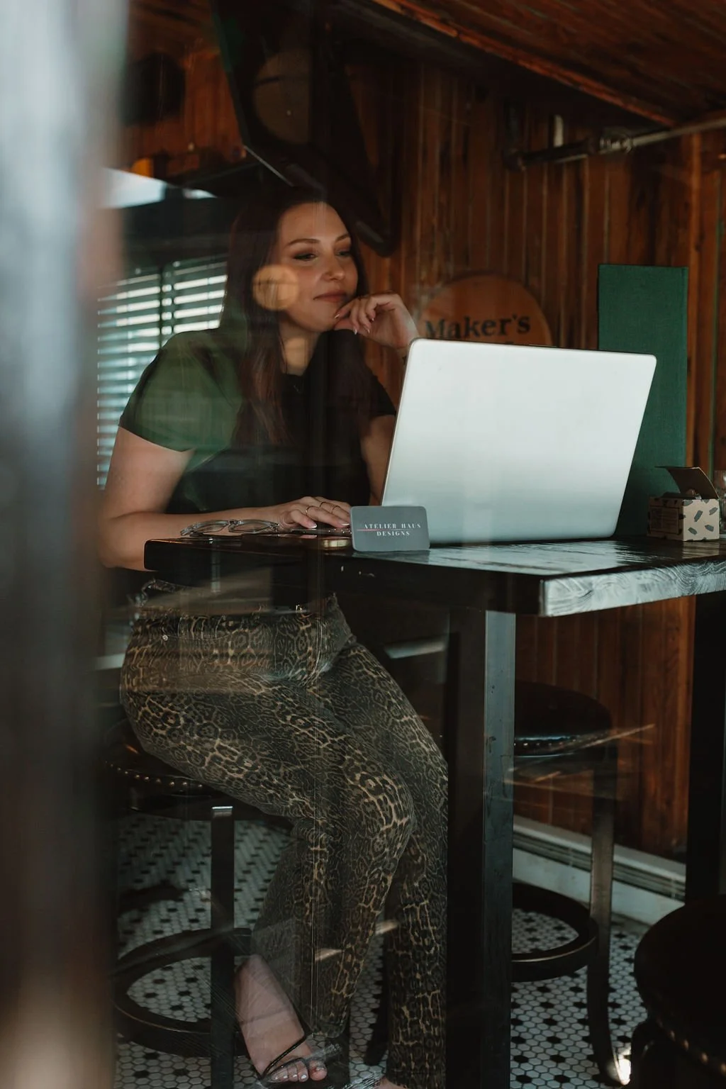 Woman sitting at a table with a laptop, wearing leopard print pants, in a wooden-paneled room, looking at the screen thoughtfully.
