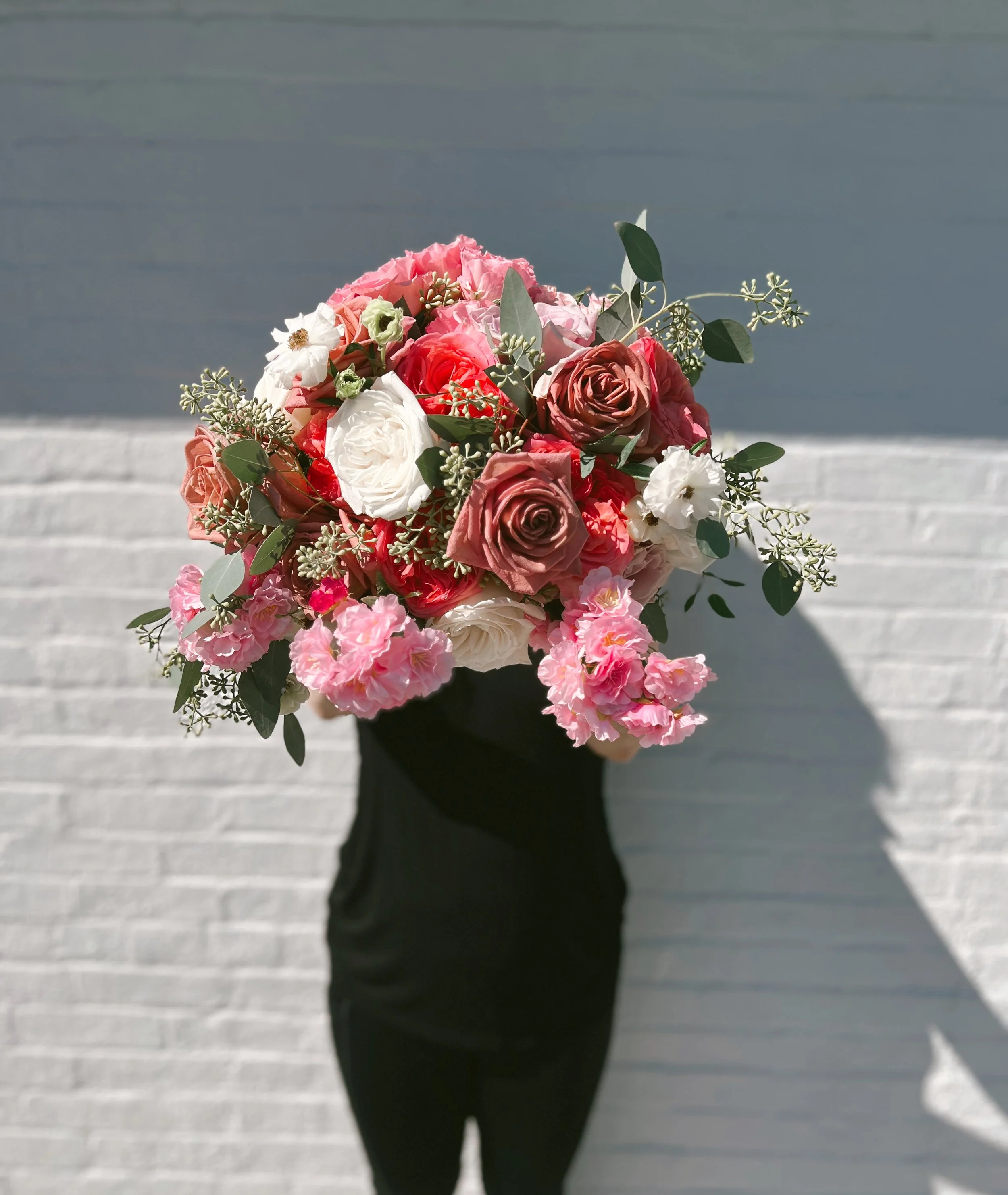 A person in black holding a large bouquet of pink, white, and blush roses with green foliage against a white brick wall.