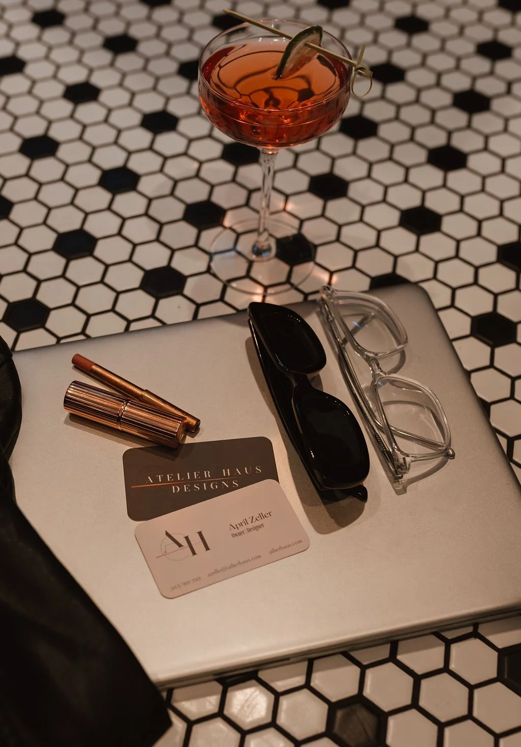 Flatlay of laptop, glasses, lipstick, and cocktail, styled by April, owner of Atelier Haus in Westminster, MD, inspiring a creative workspace for Carroll County clients