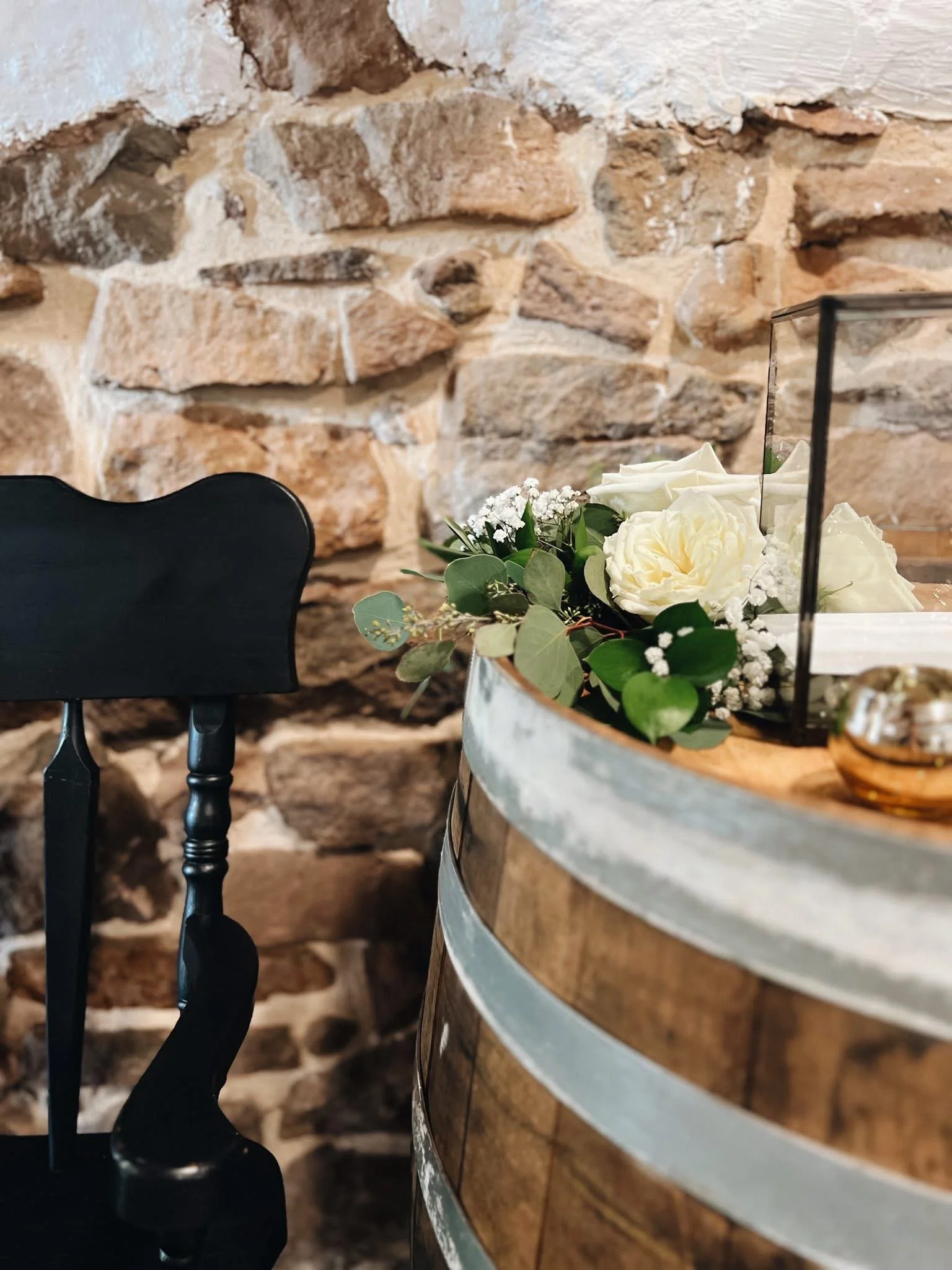 Close-up of a flower arrangement with white roses and greenery on a wooden barrel, with a black chair and stone wall in the background.
