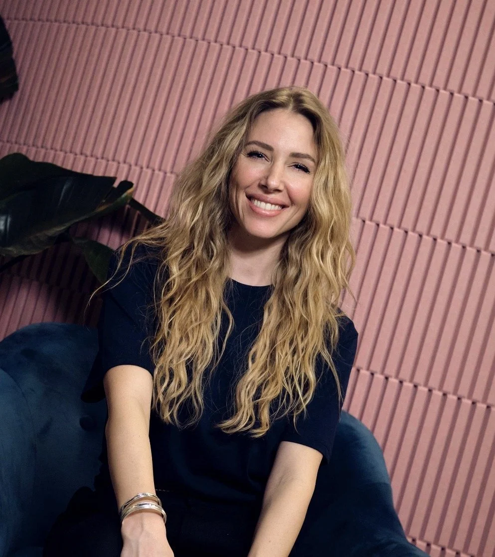 A smiling woman with long blonde curly hair, wearing a black shirt, sitting on a dark blue sofa against a pink textured wall.