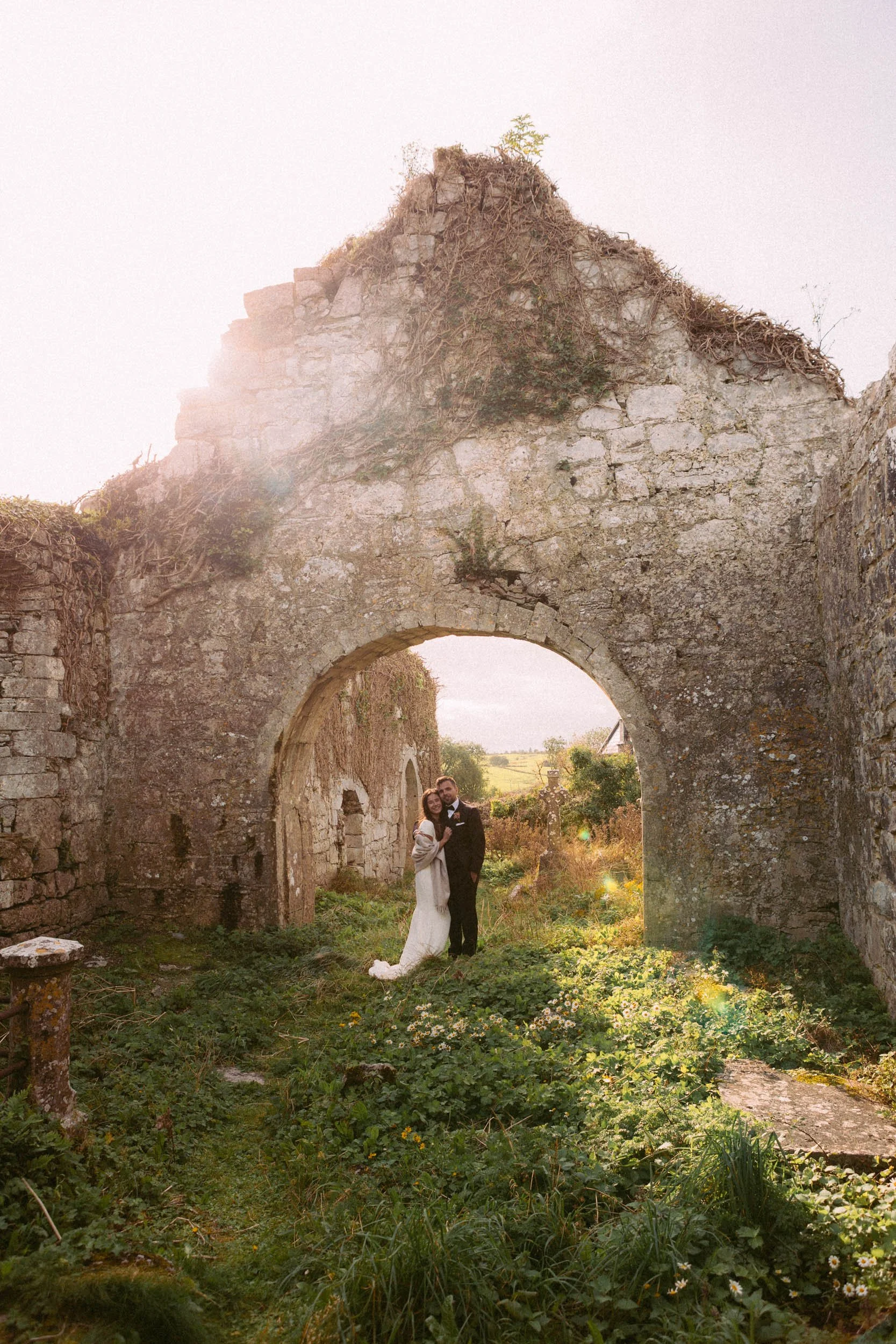 A newlywed couple in wedding attire standing inside the ruins of an old stone archway at sunset, surrounded by greenery and wildflowers.