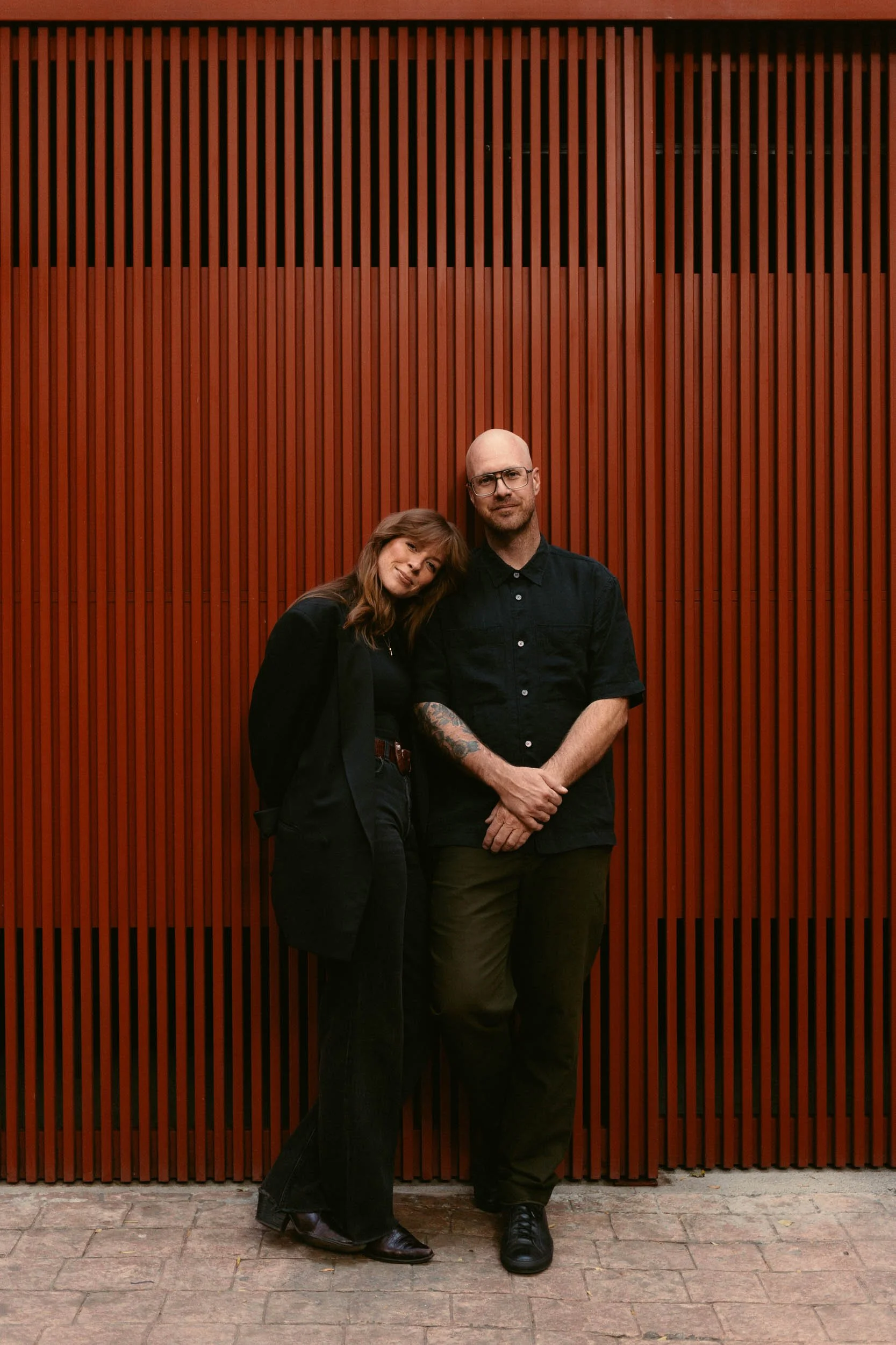 A woman and a man standing close together against a red wooden slat wall, with the woman resting her head on the man's shoulder.