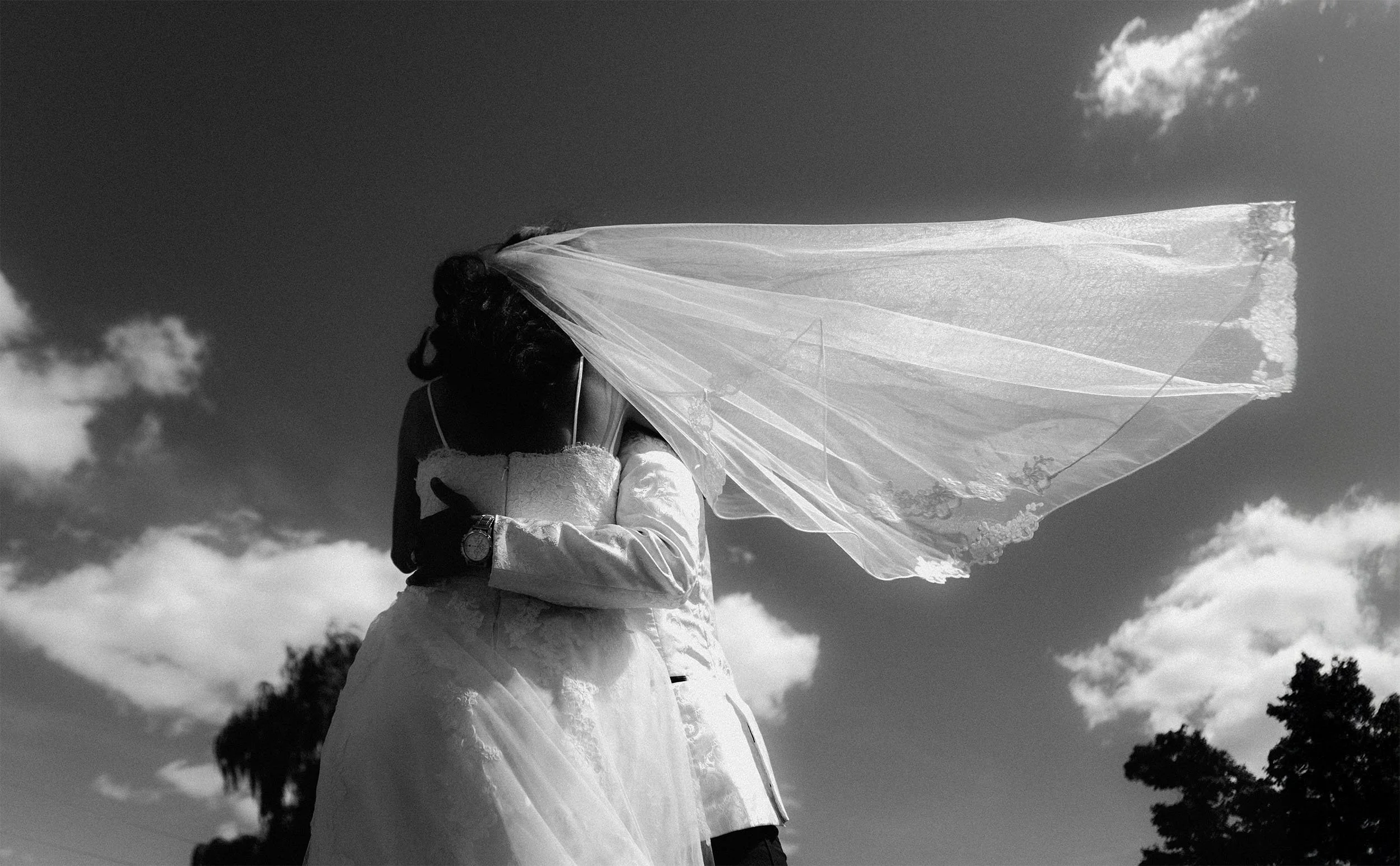 A bride and groom embrace outdoors, with the bride wearing a veil and holding a pillow, under a cloudy sky.