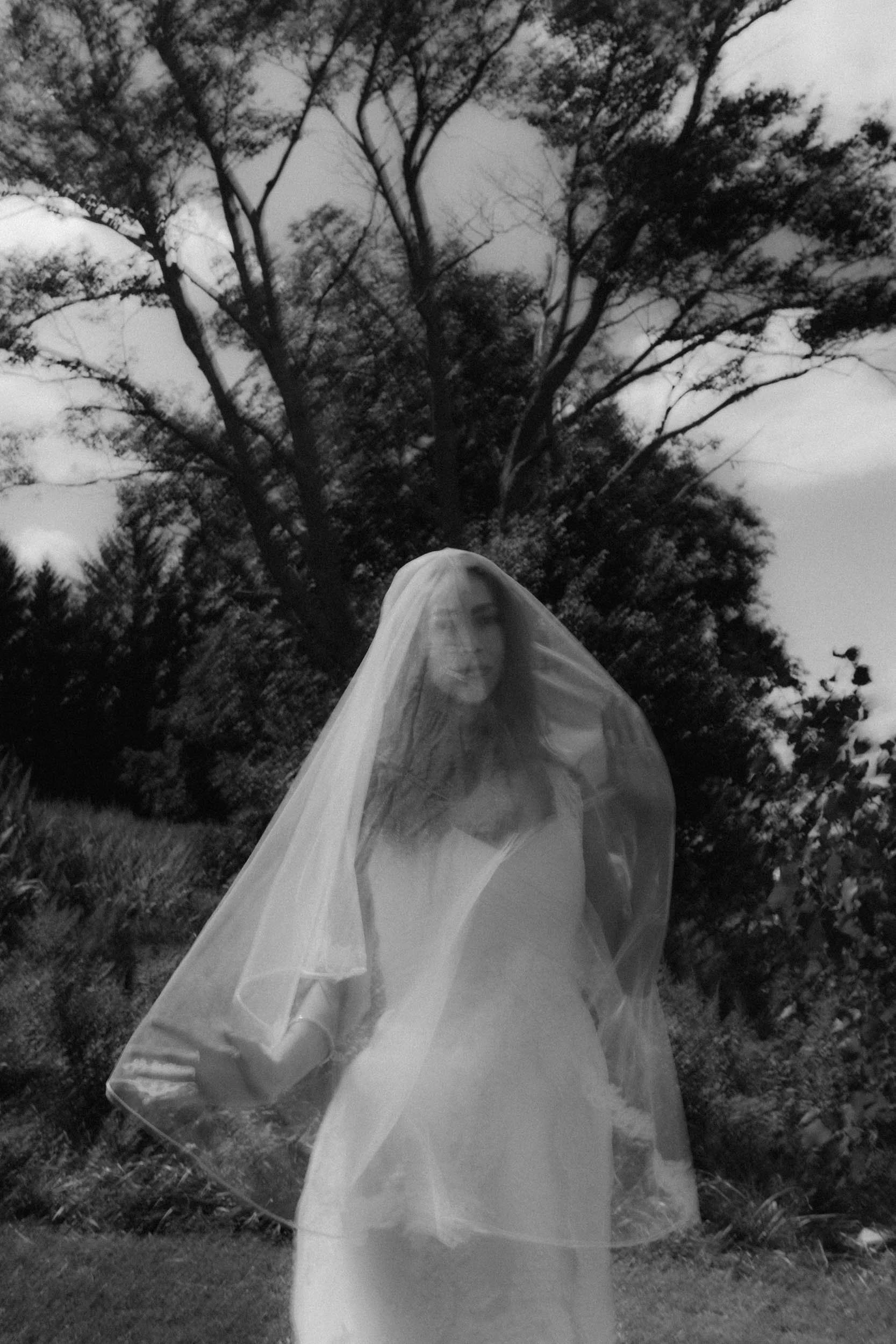 A woman in a wedding dress and veil stands outdoors in front of a large tree, with a cloudy sky overhead, in black and white.