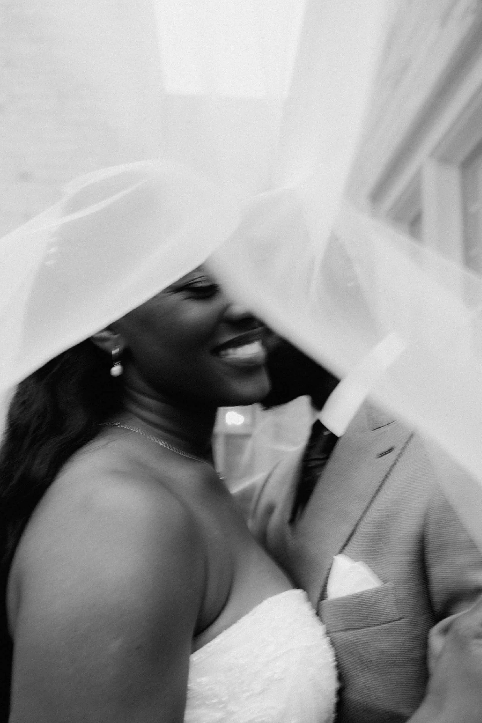 Black and white photo of a woman smiling, wearing a strapless dress and pearl earrings, with a large hat, standing close to a man in a suit.