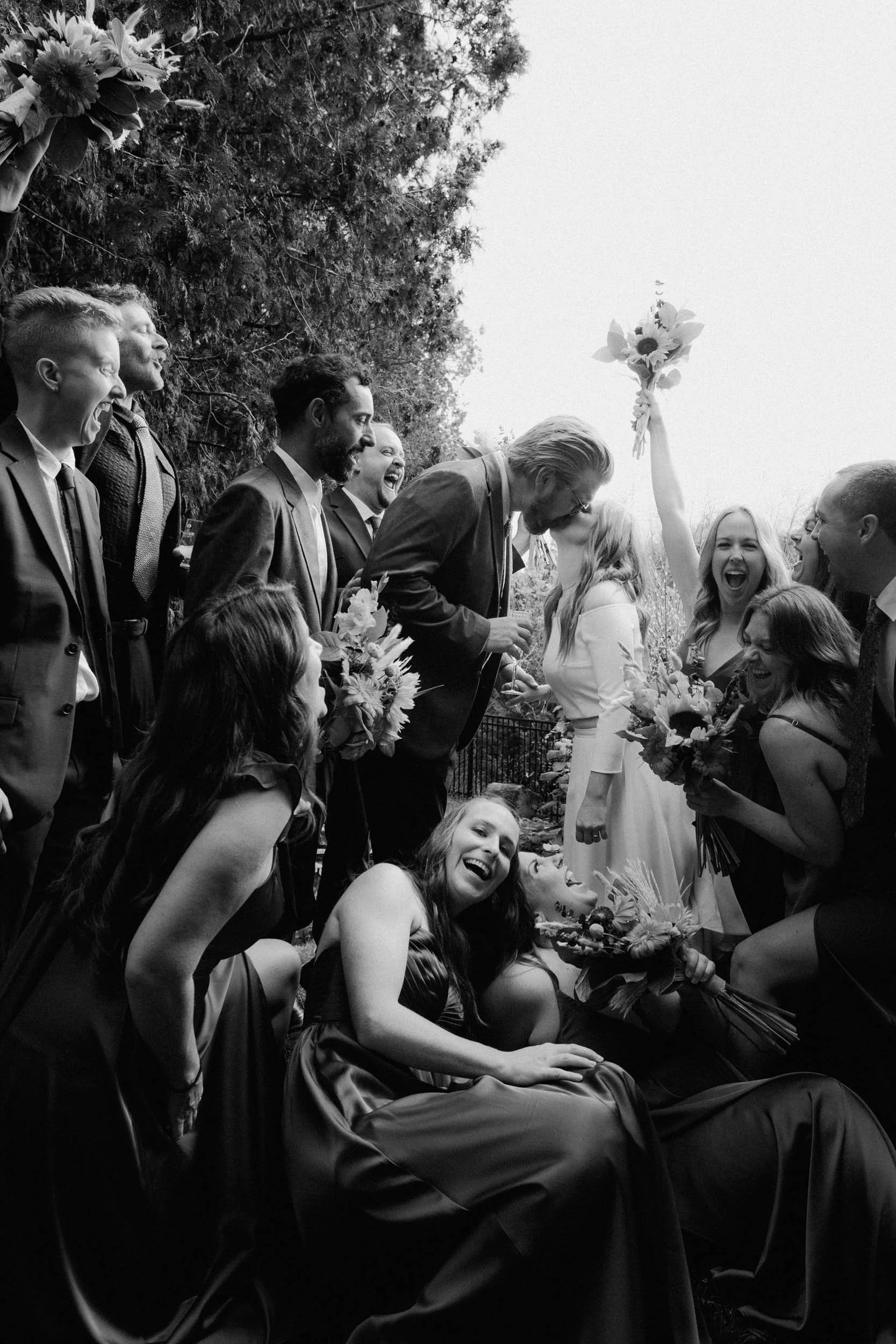 A black and white photo of a joyful wedding scene with the bride and groom kissing, surrounded by smiling guests, some holding bouquets, outdoors near trees.
