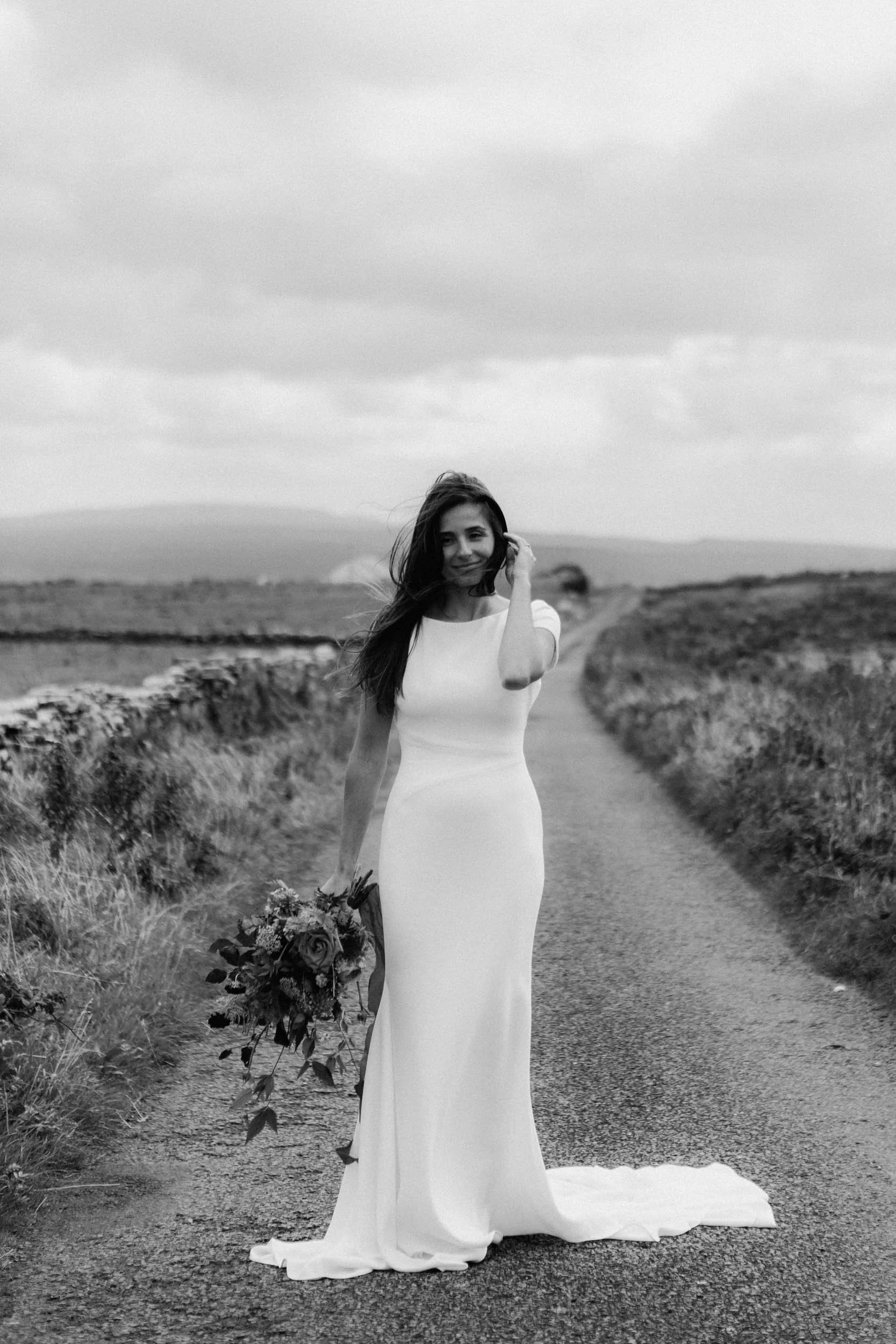 A woman in a white wedding dress standing on a rural dirt road holding a bouquet of flowers, with wind blowing through her hair, in an outdoor landscape.