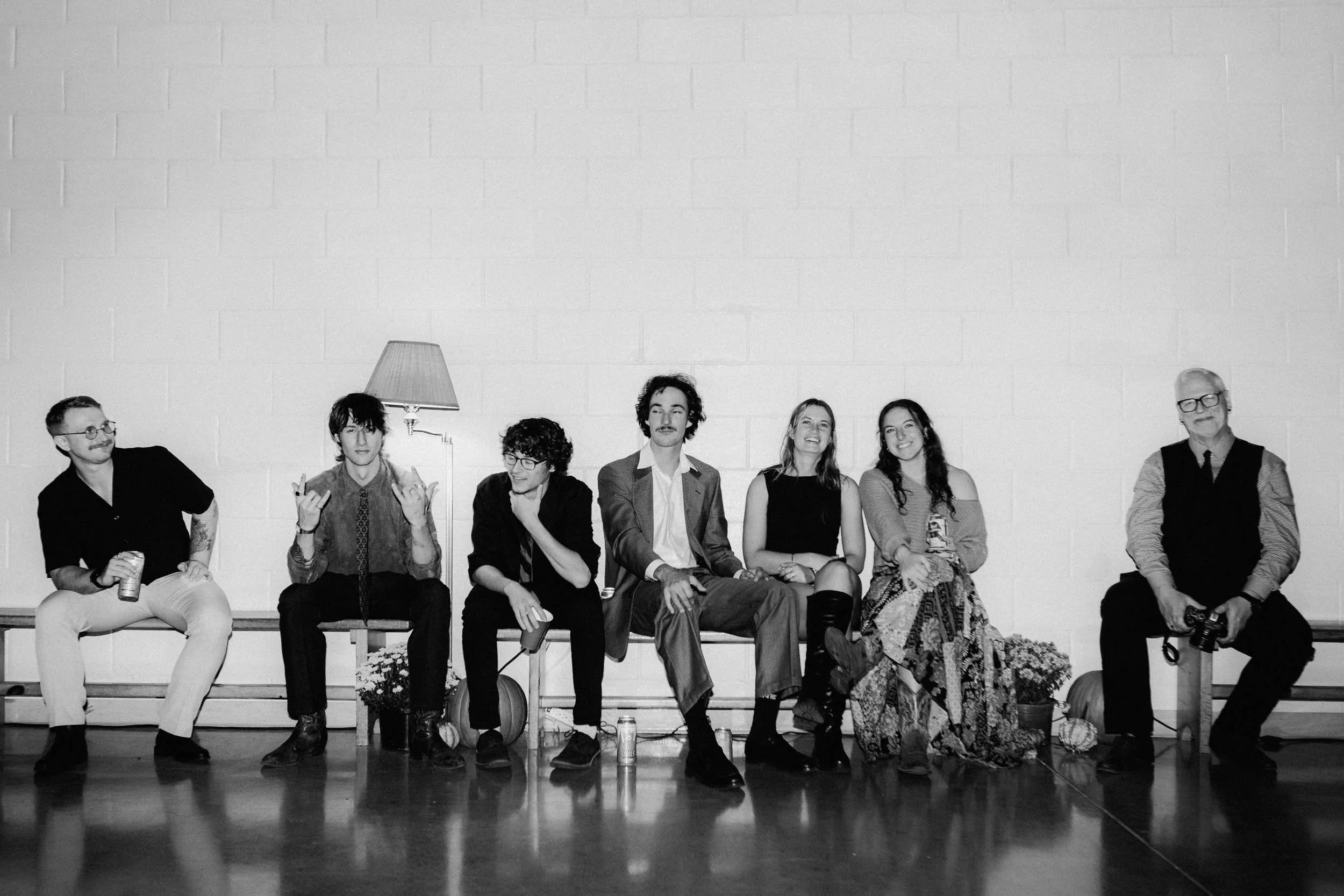 A group of seven people sitting on a bench against a white brick wall, with a lamp, plants, and pumpkins around them, in a black and white photo.