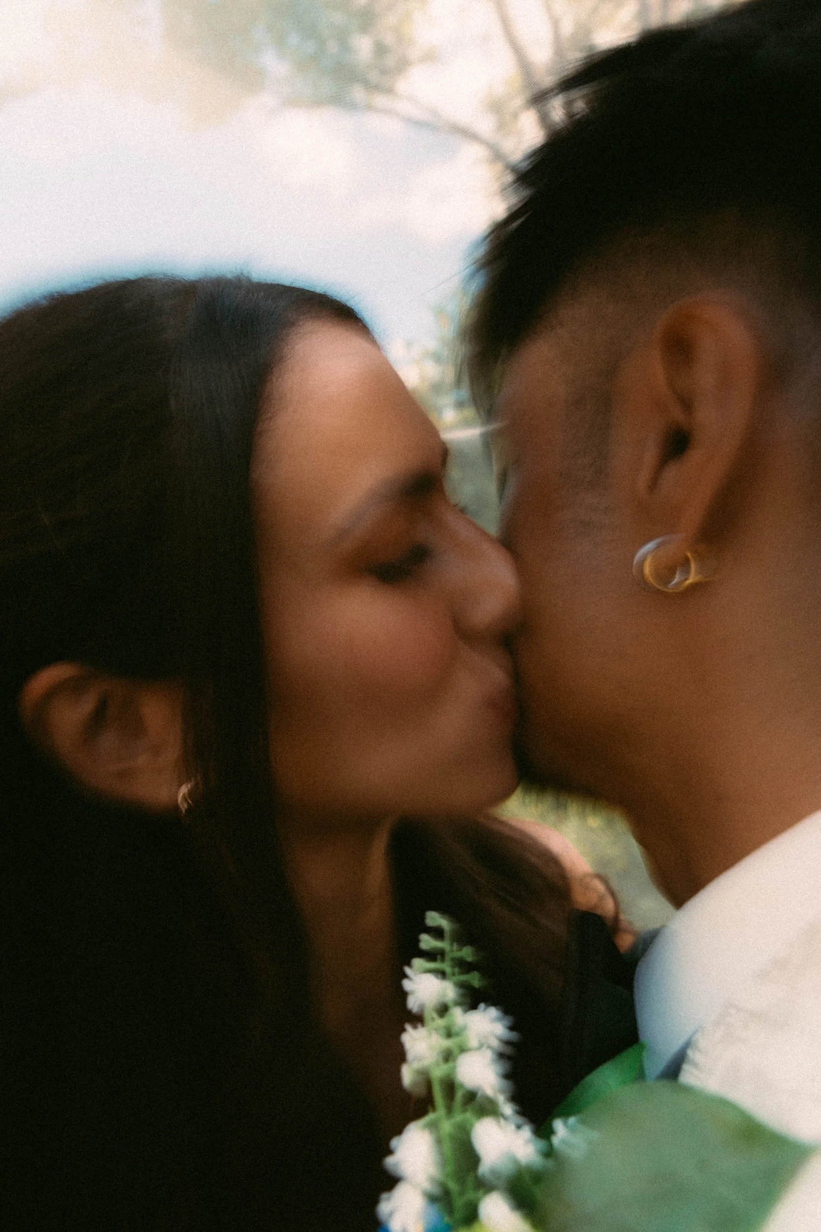 A couple kissing outdoors, with a blurred background of trees and sky.