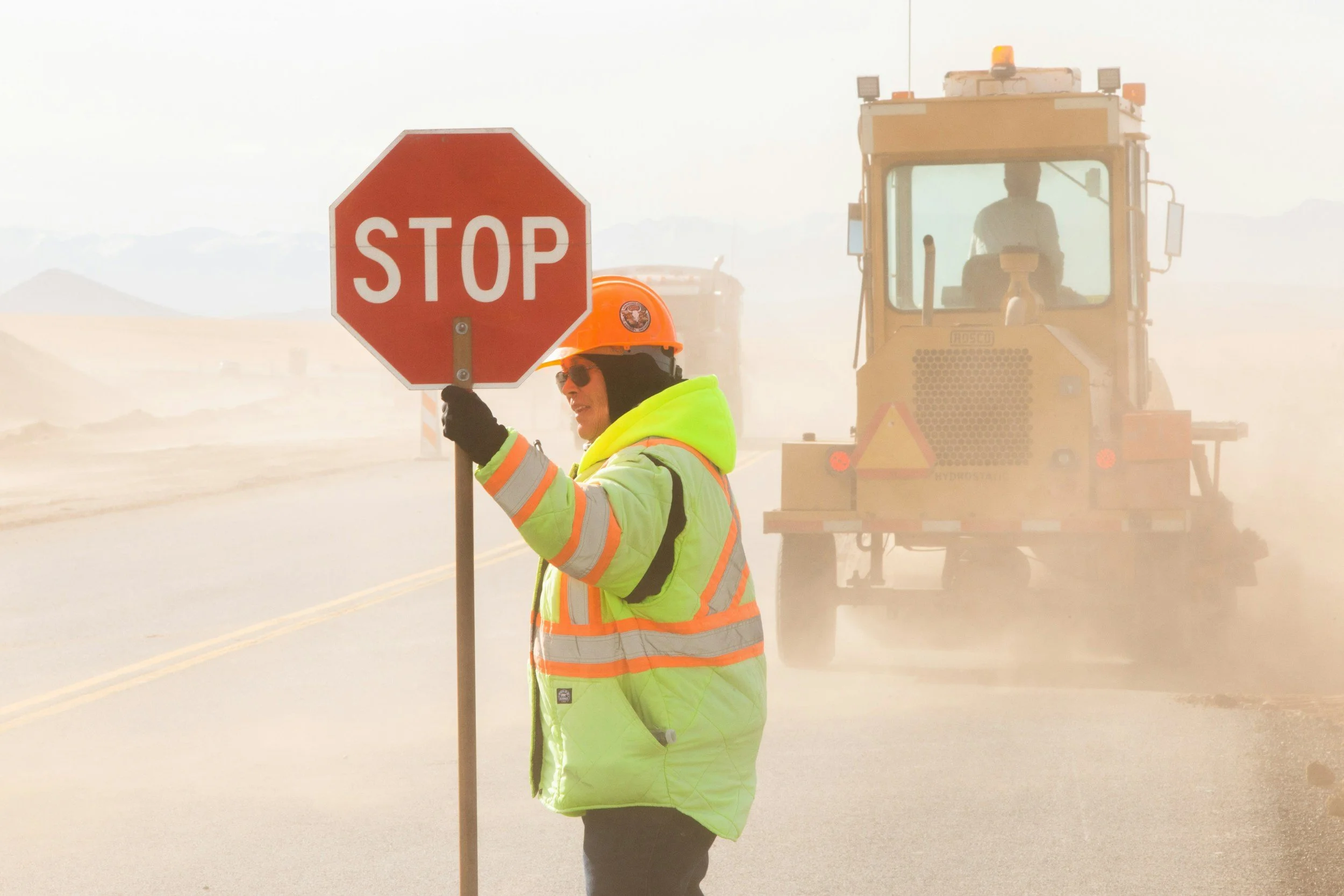 A construction worker in safety gear holding a stop sign, with a yellow construction vehicle in the background on a dusty road.