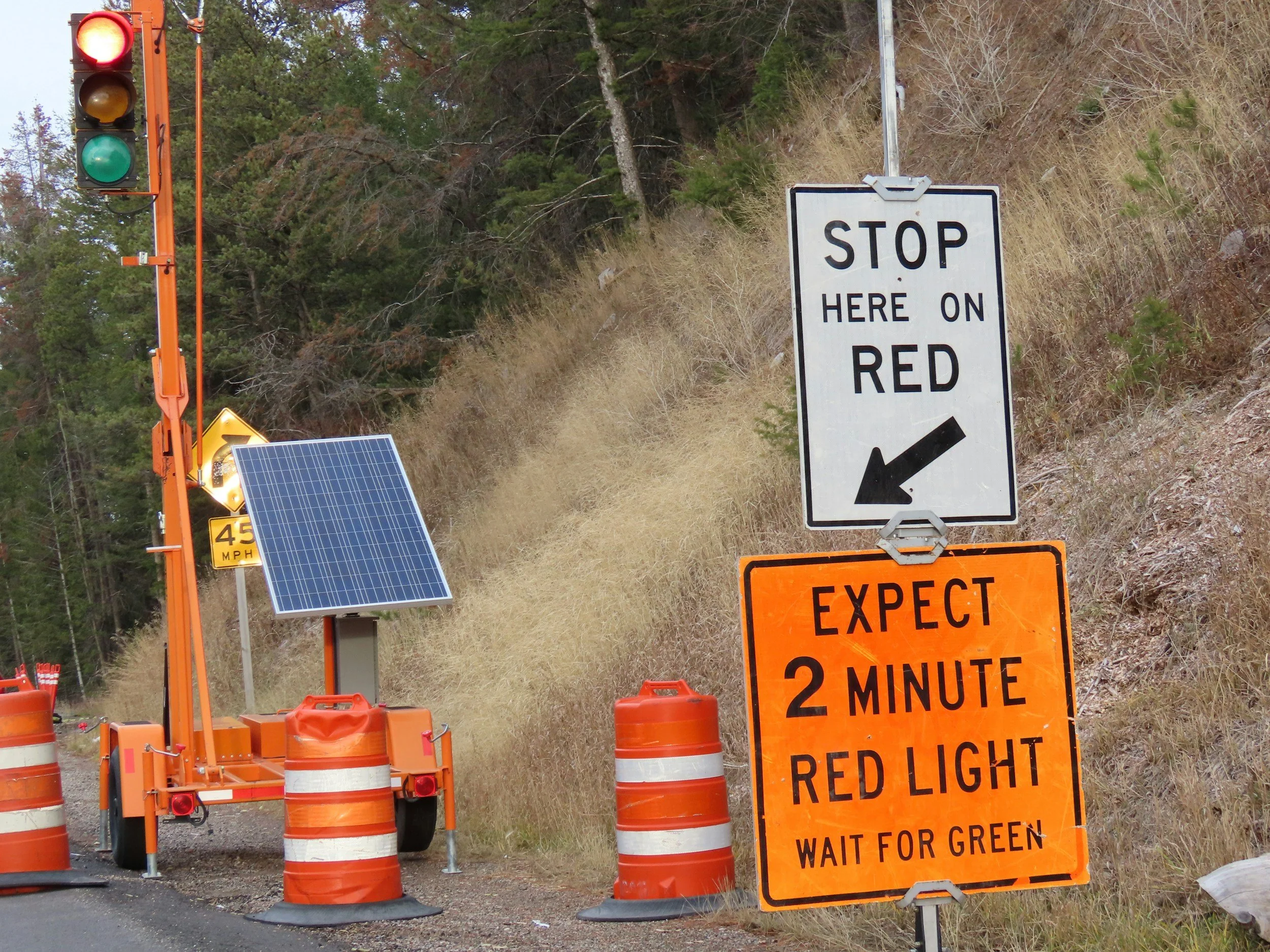 Construction zone on a road with traffic signals, orange barrels, a solar panel, and signs instructing drivers to stop at red and wait to go on green.