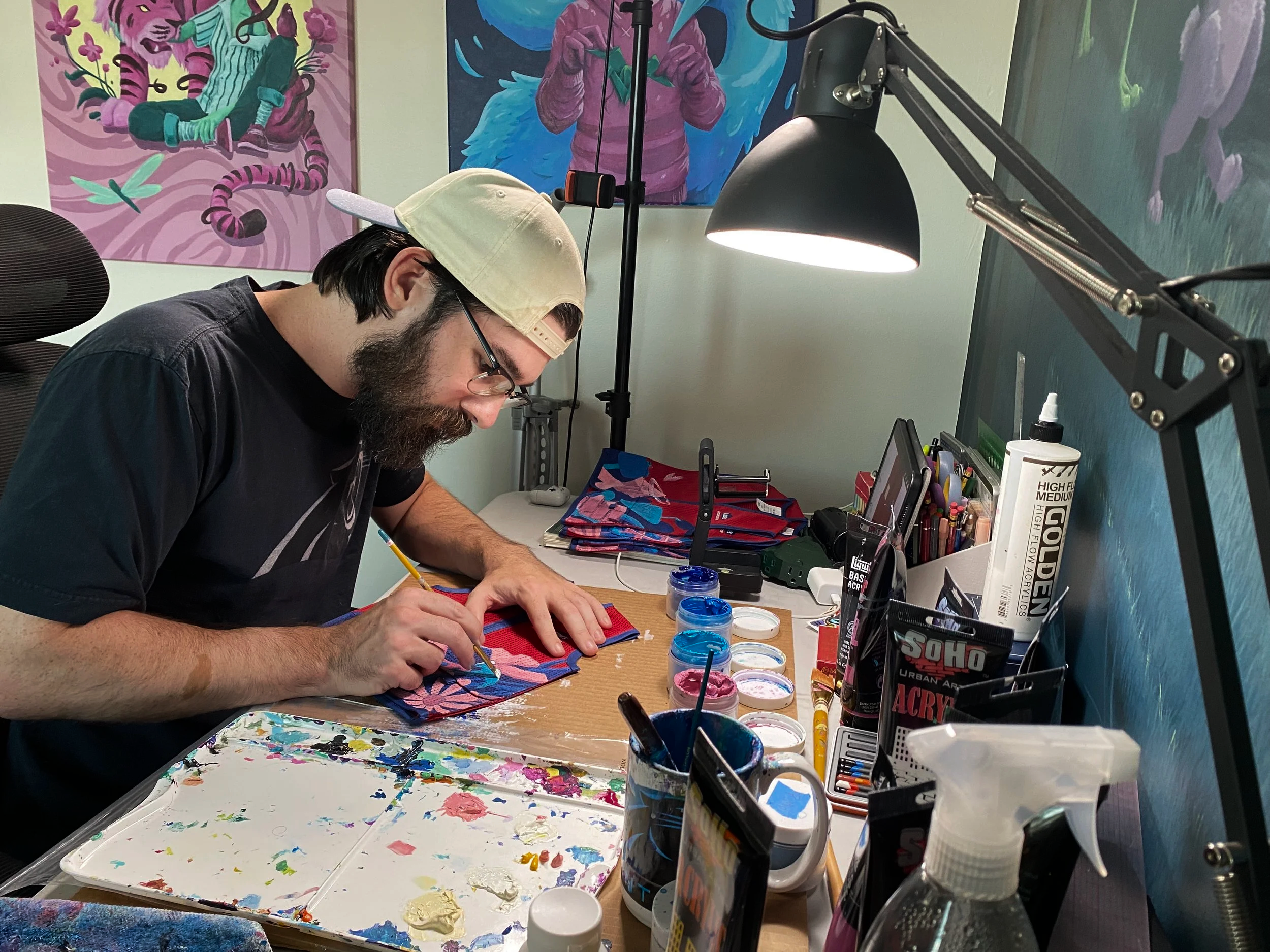 A man with glasses, a beige cap worn backwards, and a beard is painting on a small vest at a cluttered art workspace. The workspace has various art supplies, including paints, brushes, and markers, and a black desk lamp illuminating the area.