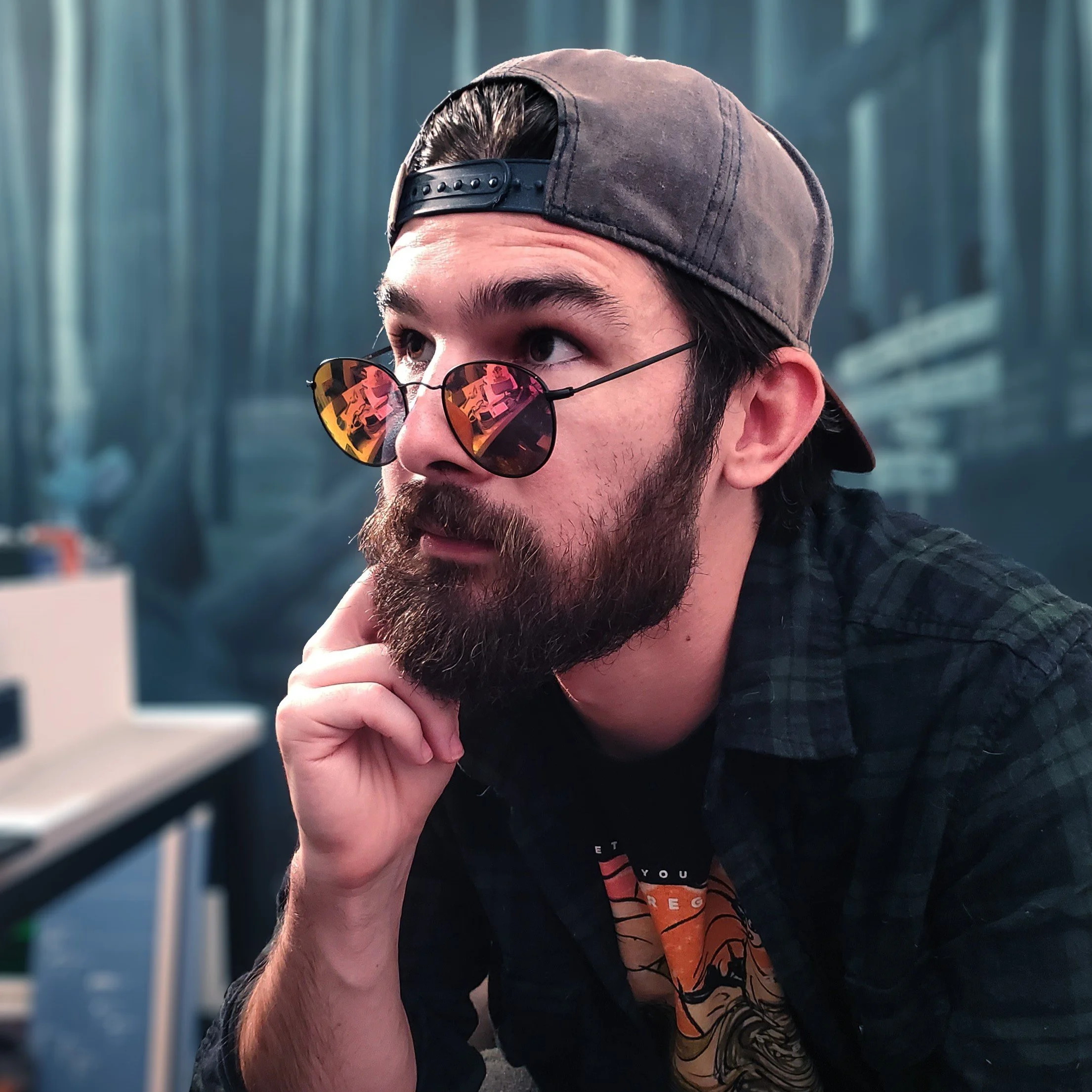 Young man with a beard wearing a backwards gray cap and reflective sunglasses, resting his chin on his hand in a room with blurred background.