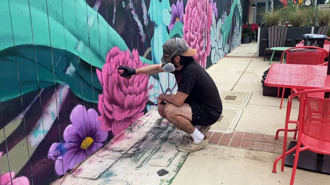 An artist crouching down on the sidewalk, spray painting a colorful mural of flowers on a fence. The person is wearing a cap, face mask, gloves, t-shirt, shorts, and sneakers.