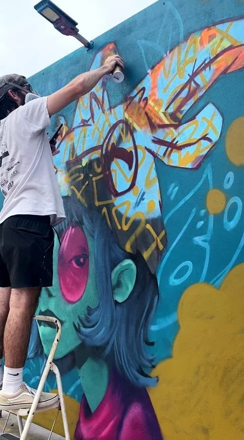 An artist is adding mural art to a large wall. The mural depicts a stylized person with blue hair, pink tinted glasses, and various colorful bubble elements.