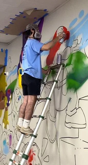 Artist standing on a ladder painting a large mural on a wall. This section of the mural features a stylized rendition of the Nintendo character Mario