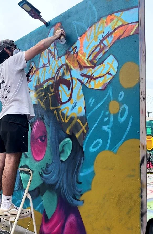 An artist is adding mural art to a large wall. The mural depicts a stylized person with blue hair, pink tinted glasses, and various colorful bubble elements.