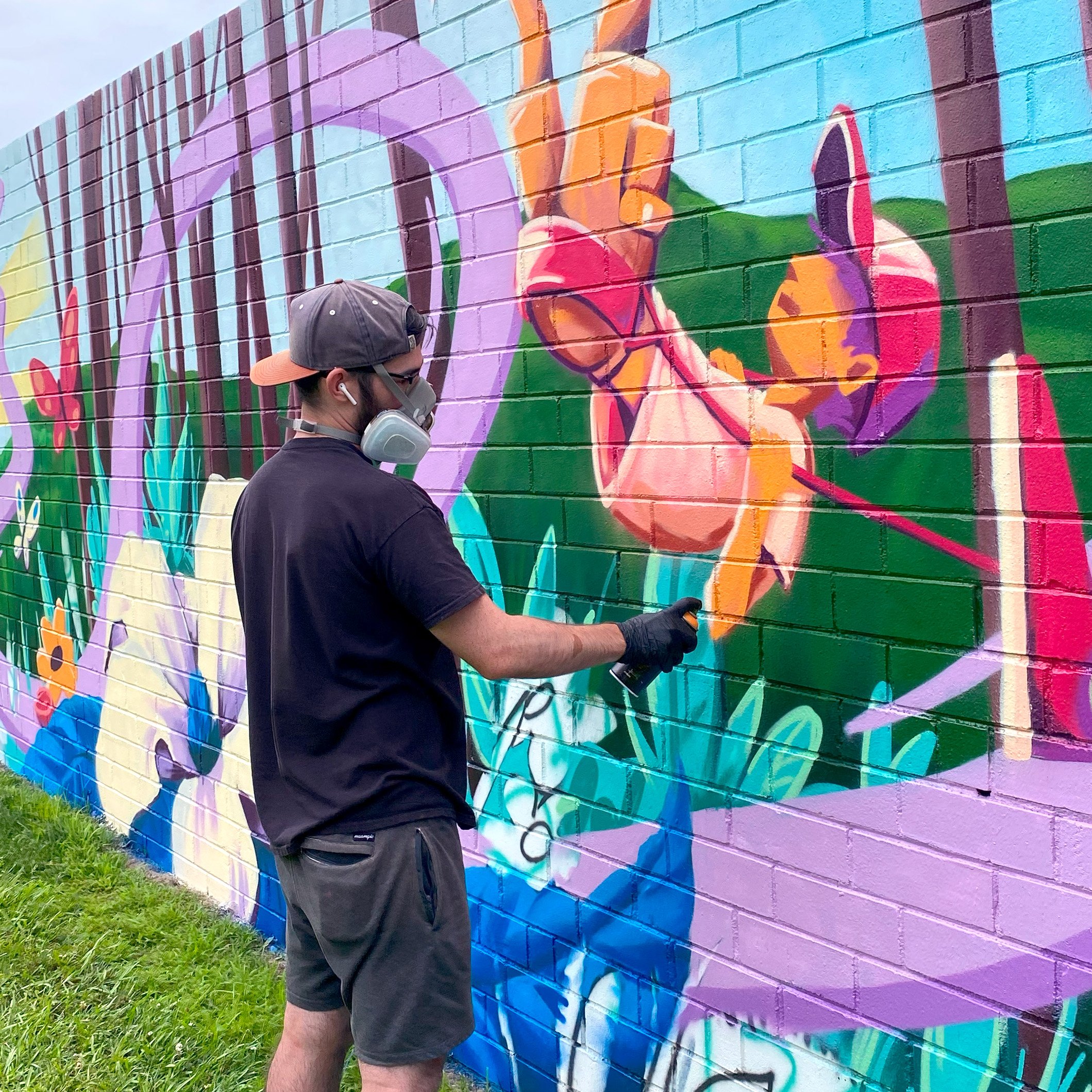 An artist spray painting a colorful mural on a brick wall. The mural features a colorful kid on a swing surrounded by vibrant flowers and foliage.