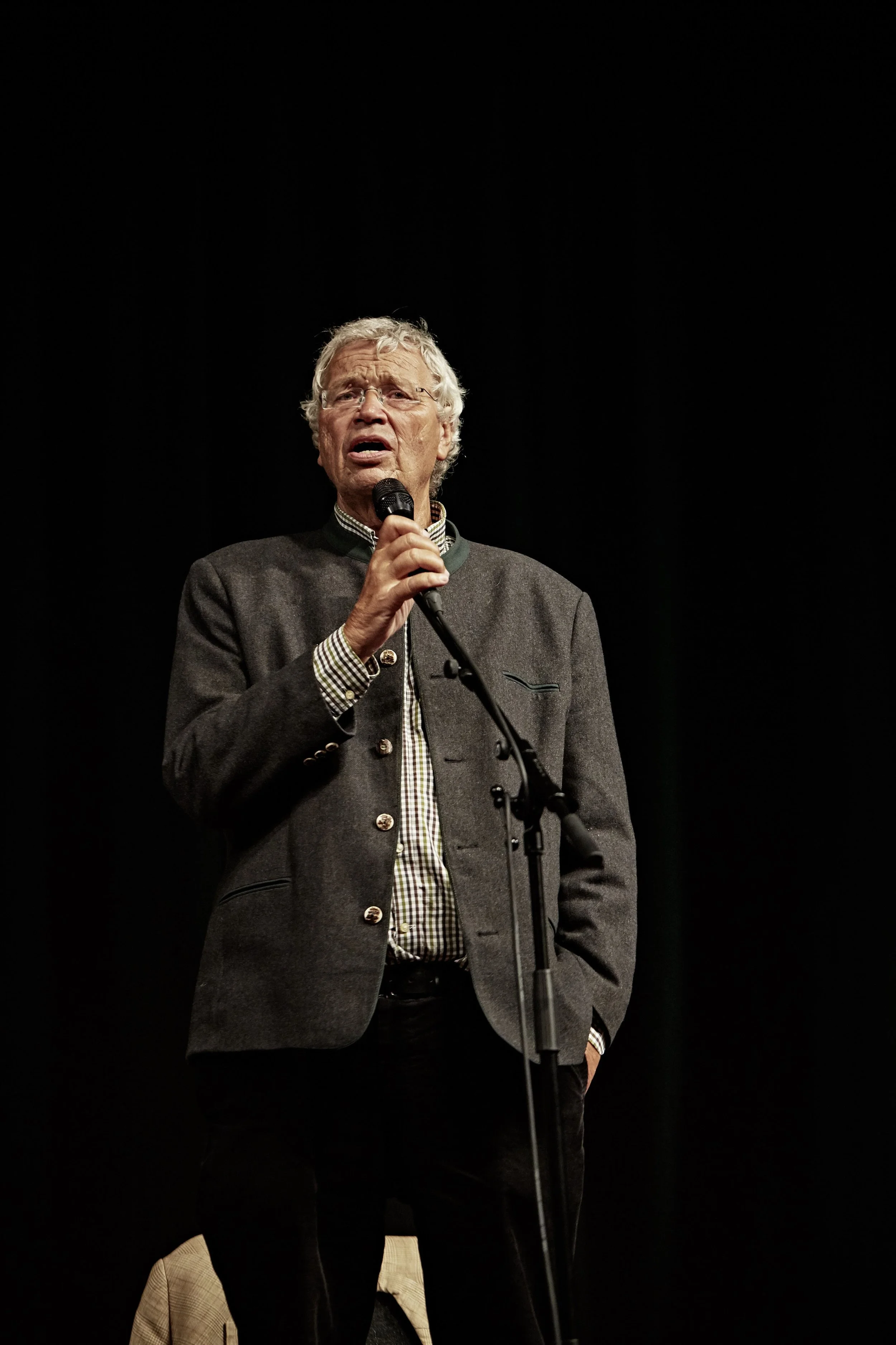 Older man wearing glasses and a gray jacket speaks into a microphone on a dark stage.