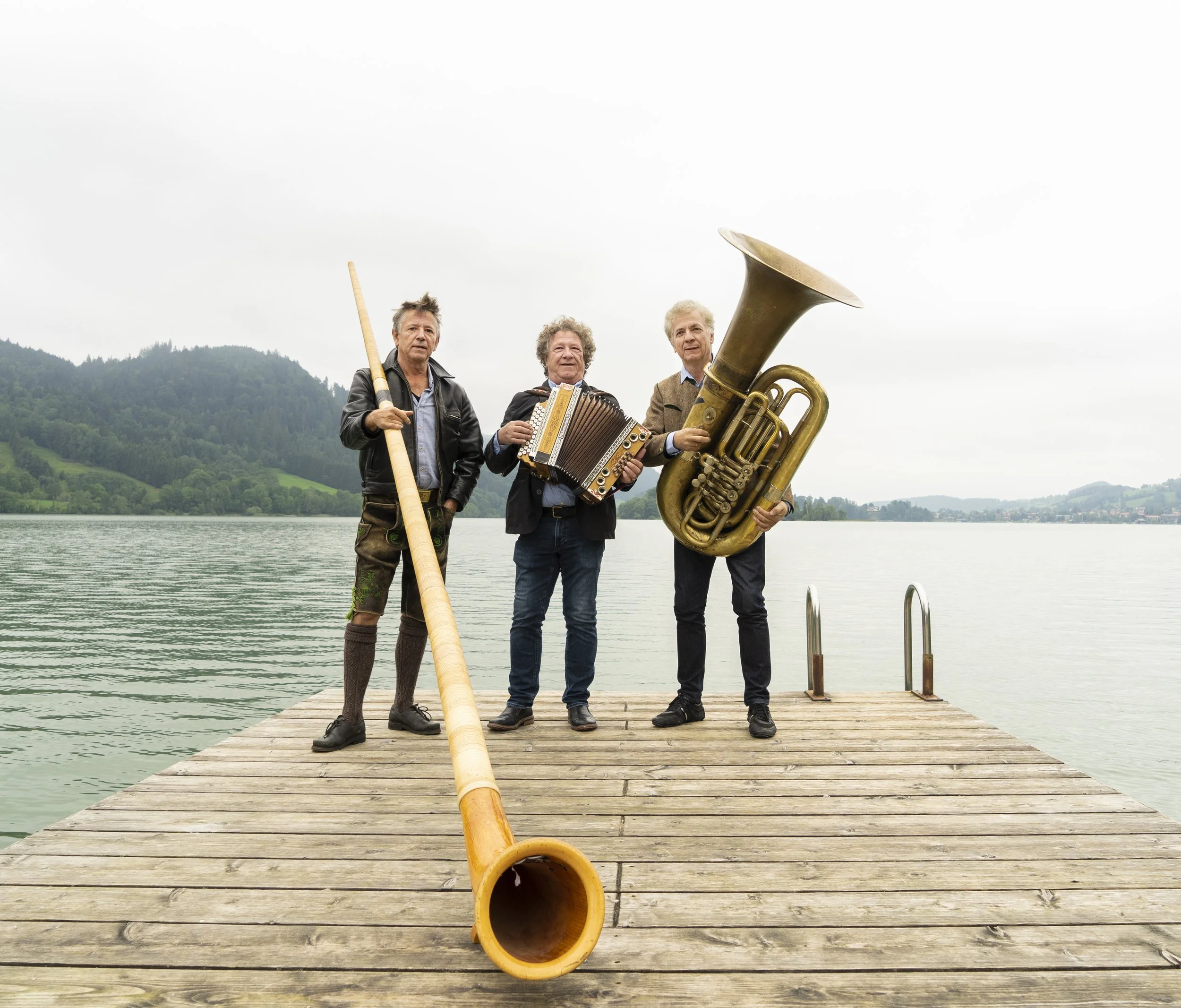 Three musicians stand on a lakeside dock with an alphorn, accordion, and large brass tuba against a mountain lake landscape.