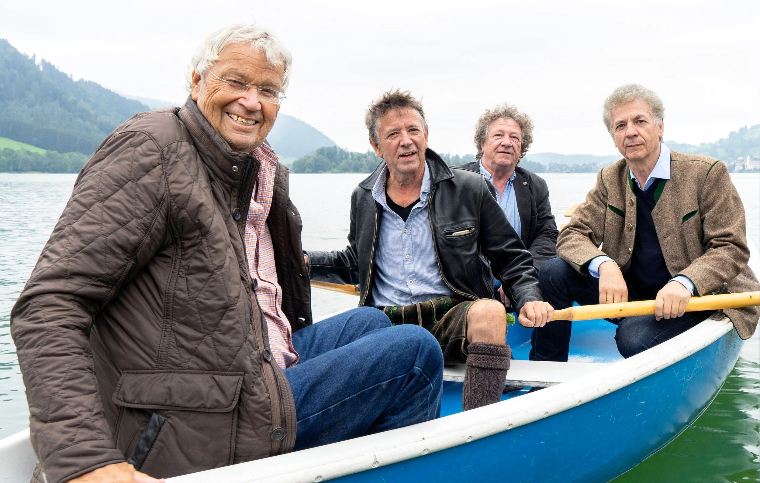 Four older men sitting in a blue rowboat on a mountain lake, with forested hills in the background.