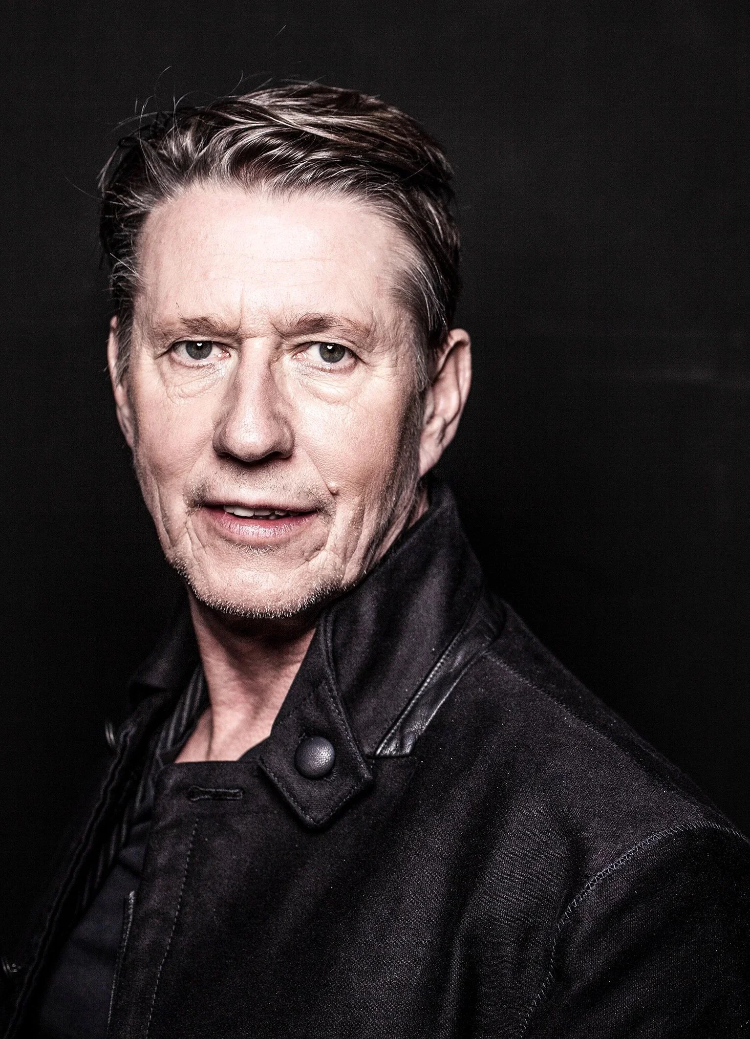 Close-up portrait of a middle-aged man with swept-back hair wearing a black jacket against a dark background