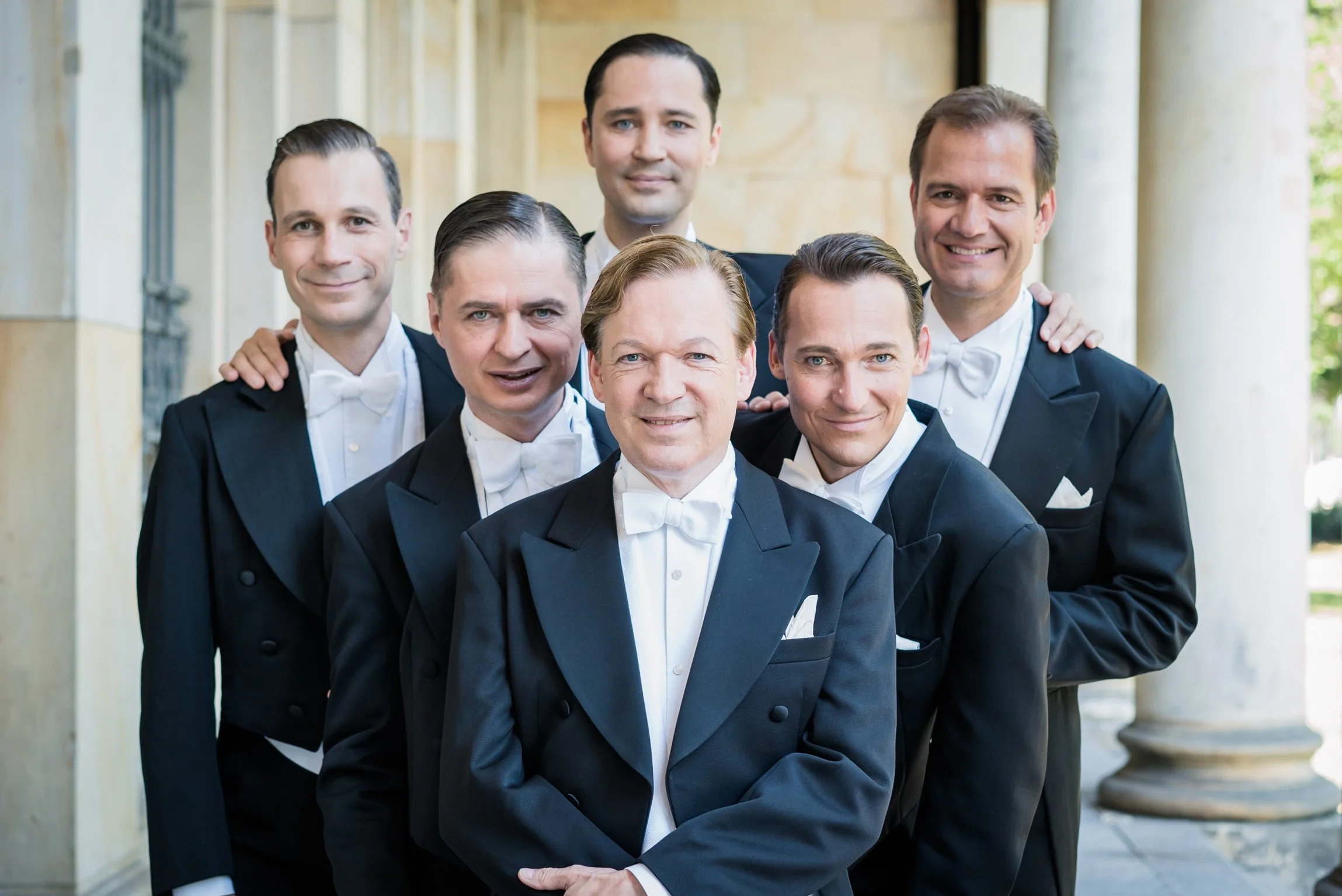 Six men in formal black tuxedos and white bow ties posing together in a bright classical setting