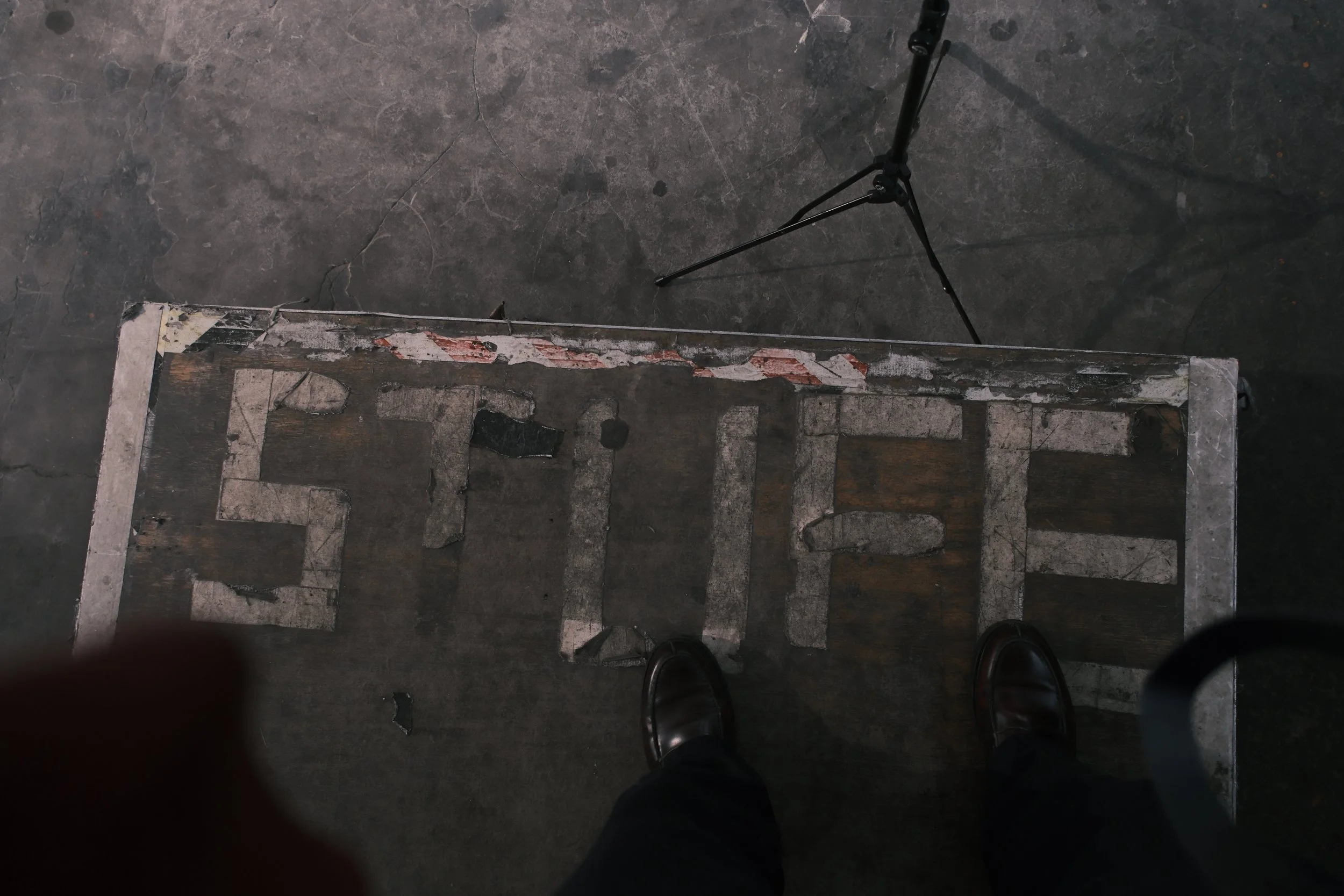 Top-down view of a worn, weathered wooden platform with peeling paint, a black tripod, and polished shoes visible on a concrete floor