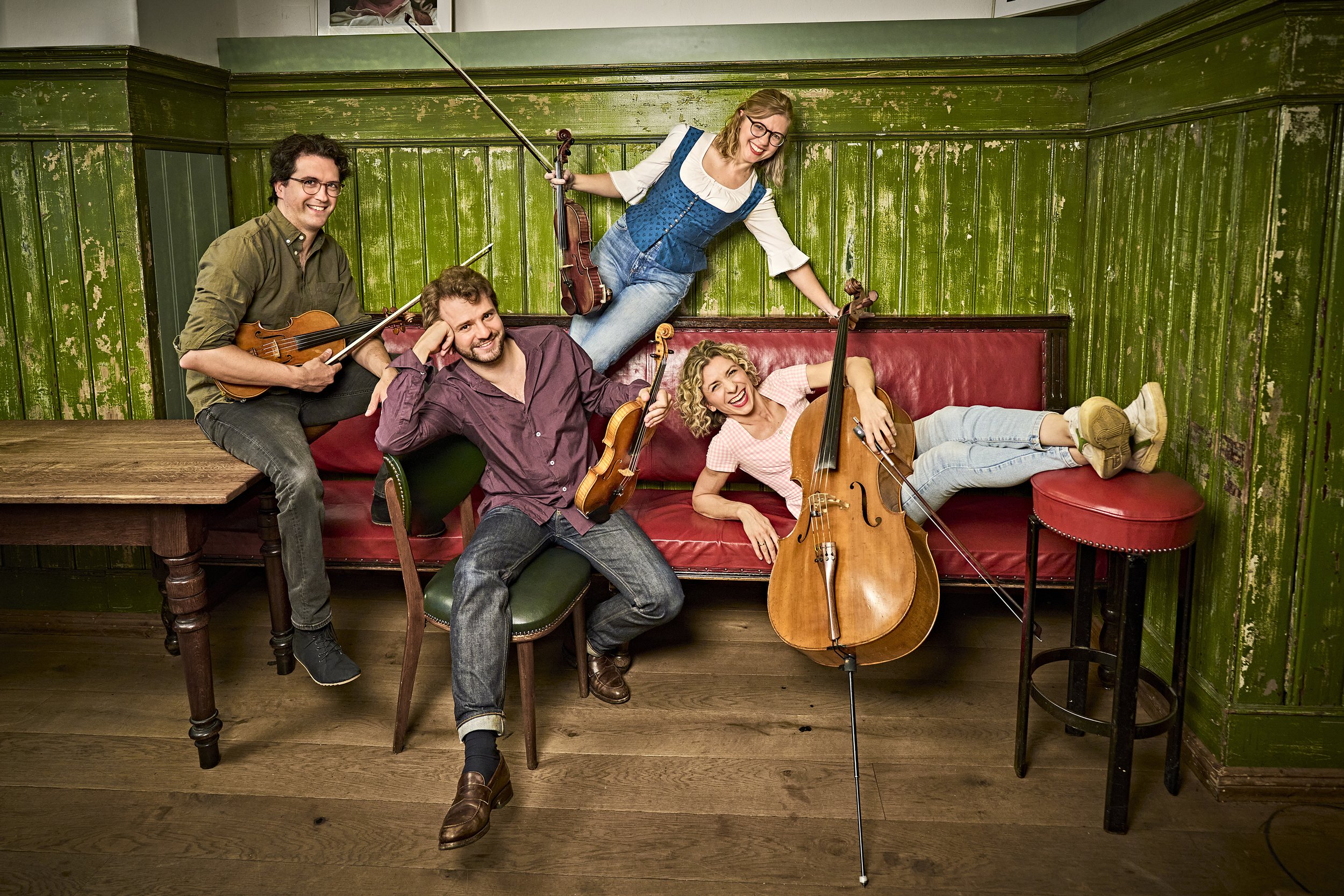 Four smiling musicians posing with violins and a cello in a rustic green-paneled room