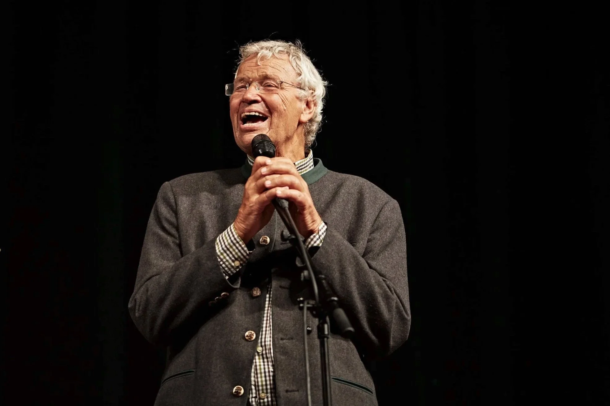 Older man laughing while speaking into a microphone on a dark stage