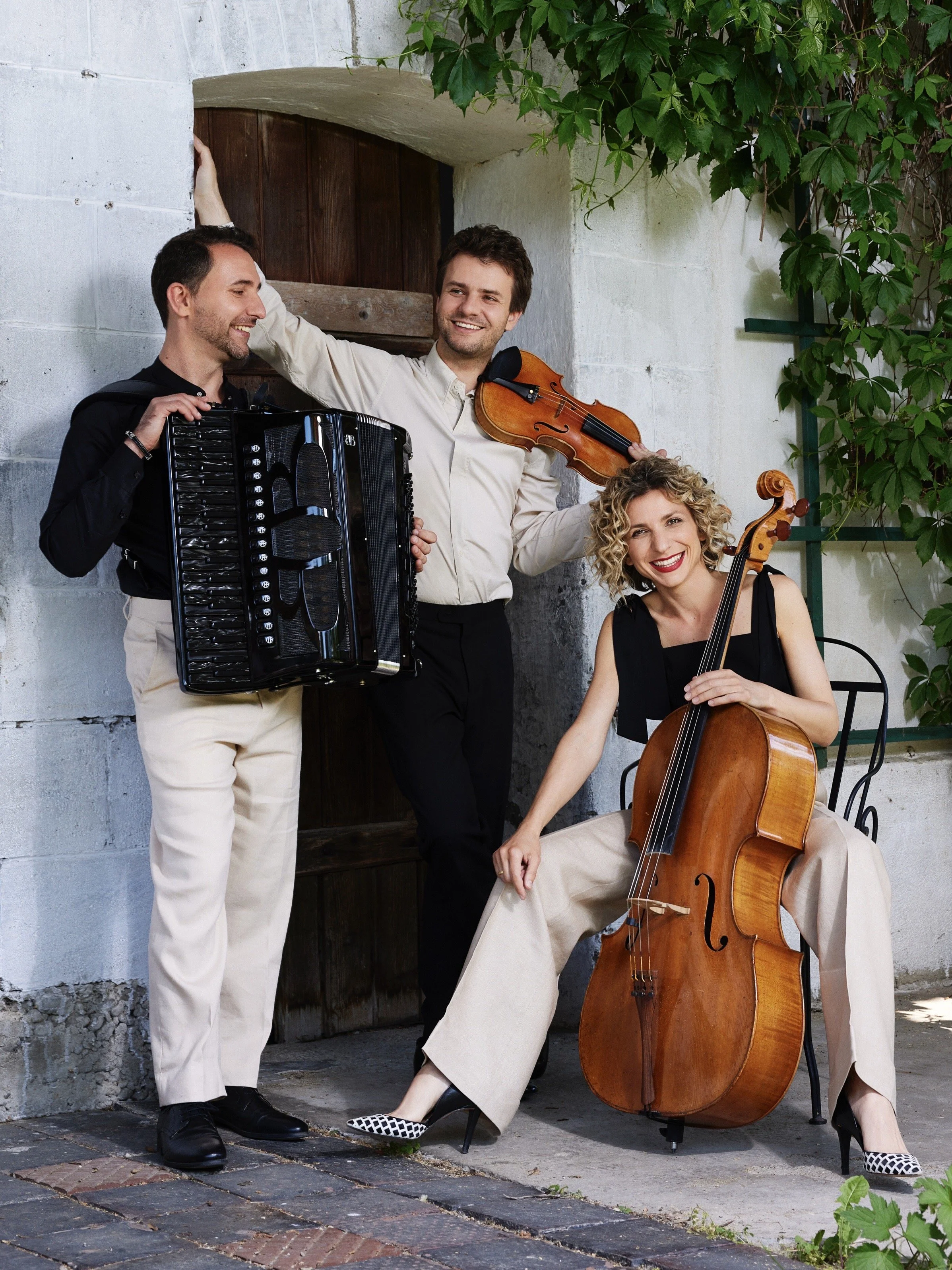 Musicians posing outdoors with an accordion, violin, and cello beside a rustic doorway covered in greenery